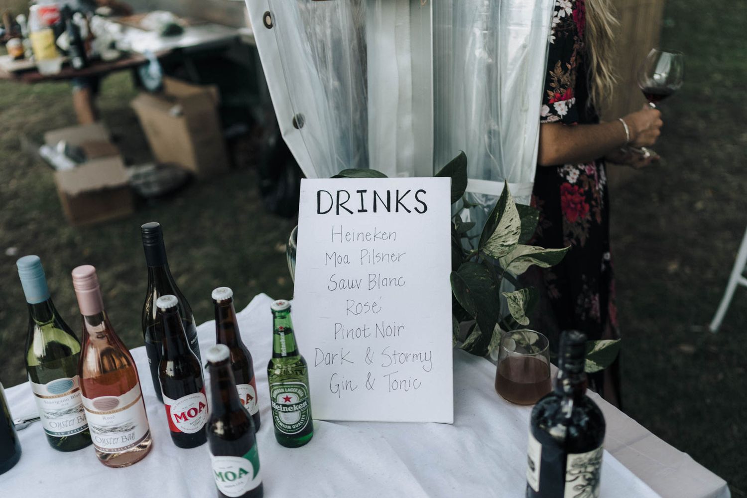An assortment of wine and beer bottles arranged outdoors with handwritten signs for drinks service.