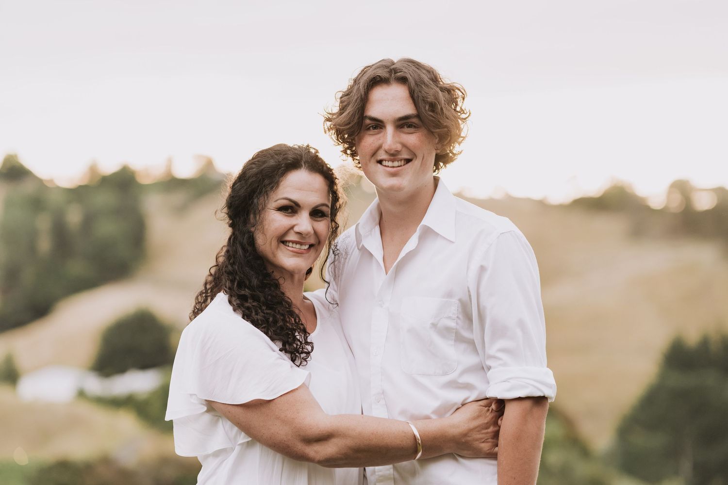 A couple in white outfits embrace while smiling outdoors against a scenic hillside backdrop during golden hour.