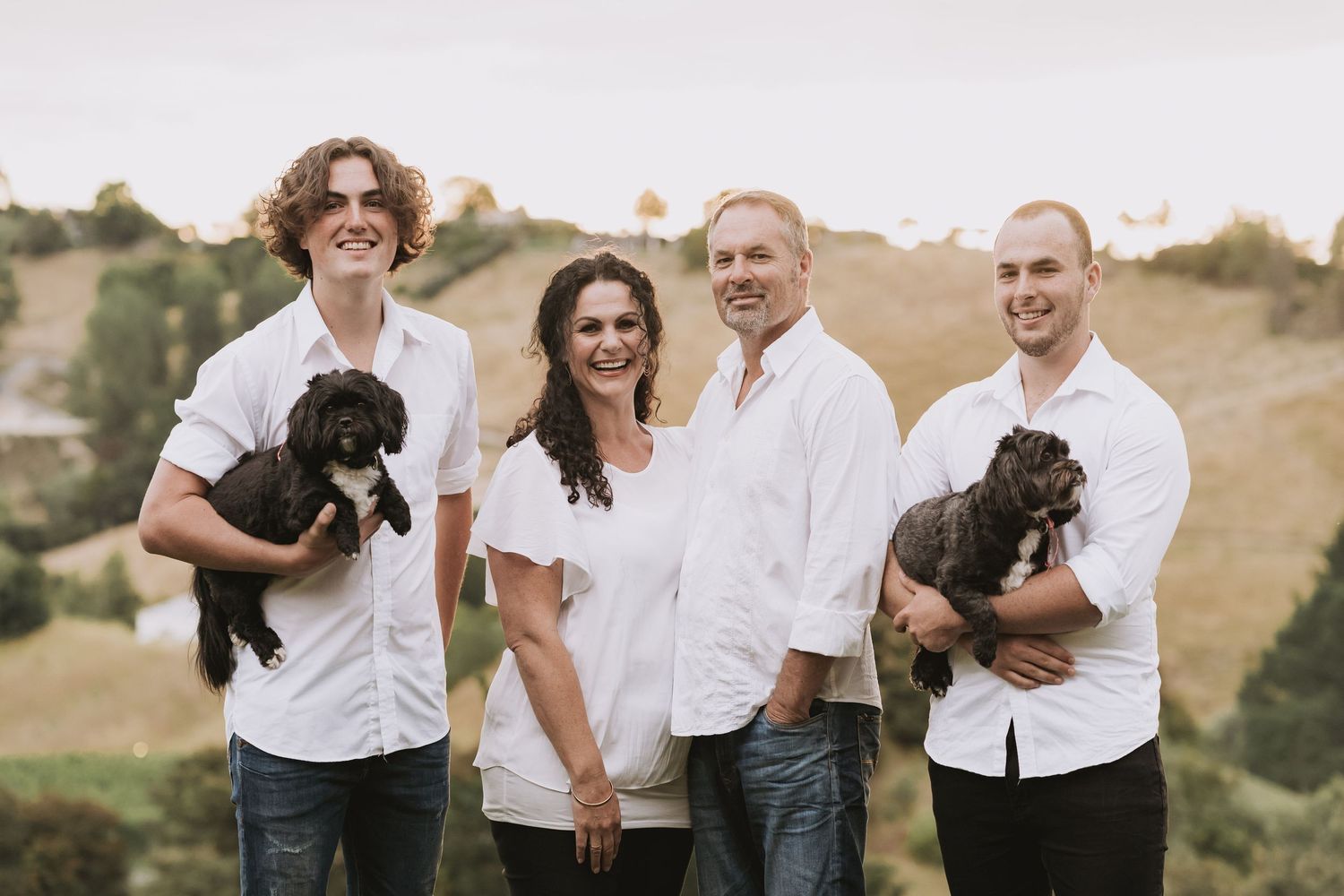 Group in white shirts stands together outdoors on rolling hills holding two black puppies while smiling at camera.