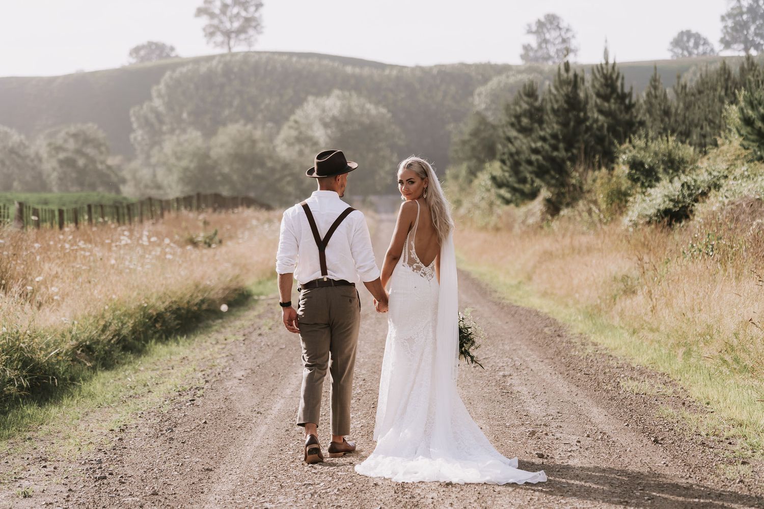 A couple walks hand in hand down a rustic dirt road surrounded by golden grass and rolling hills on their wedding day.