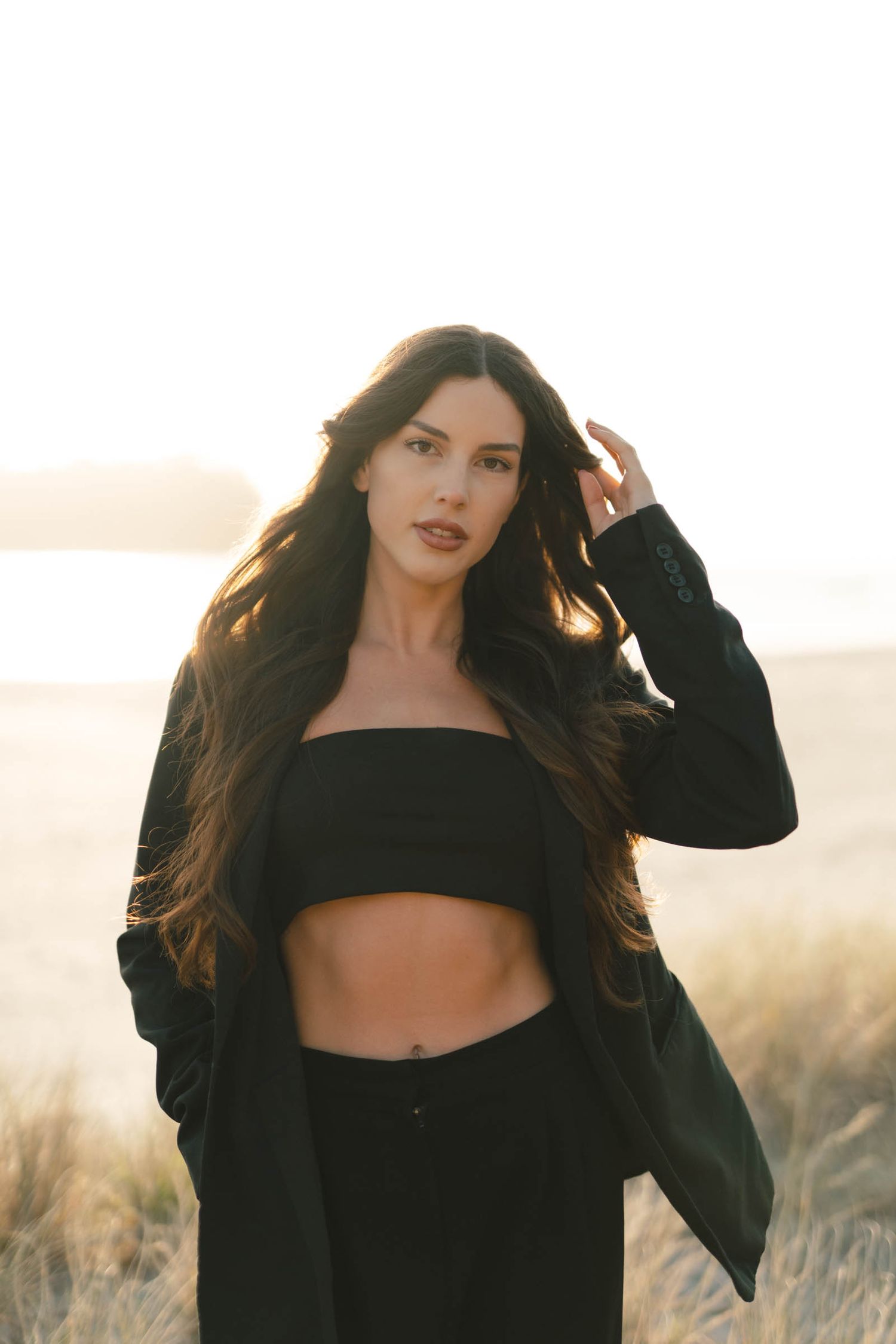 Woman in black crop top and blazer poses against sunset in grassy field.