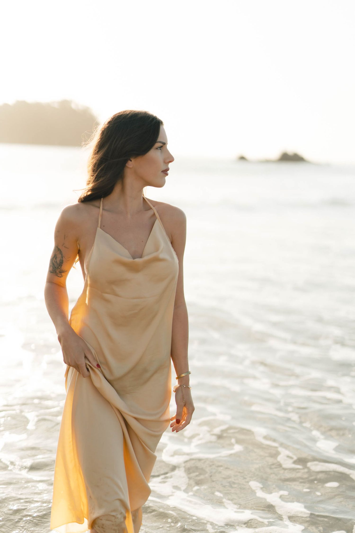Figure in silk dress stands in ocean waves during sunset.