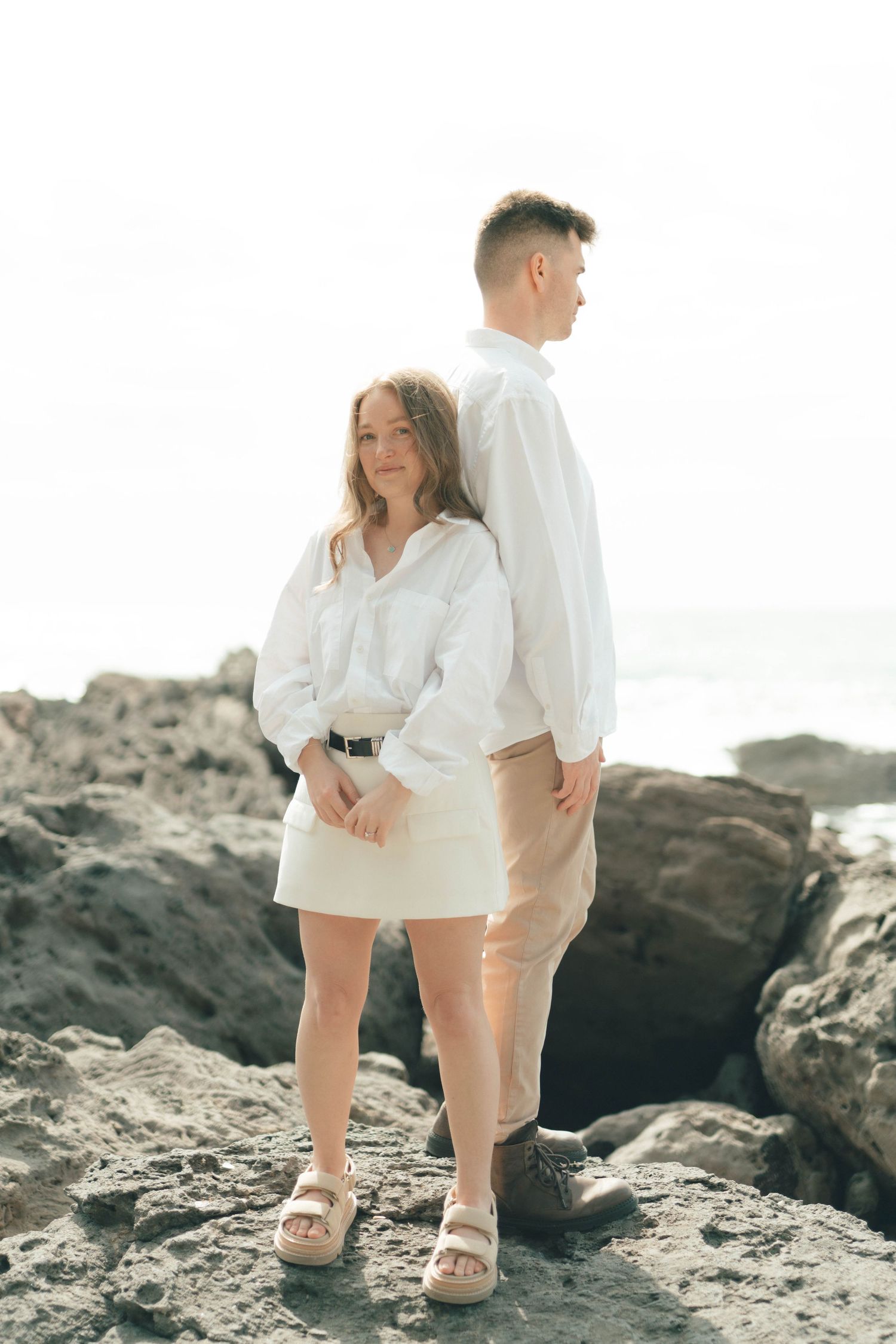 Two people in white outfits walking along rocky coastal terrain with waves nearby.