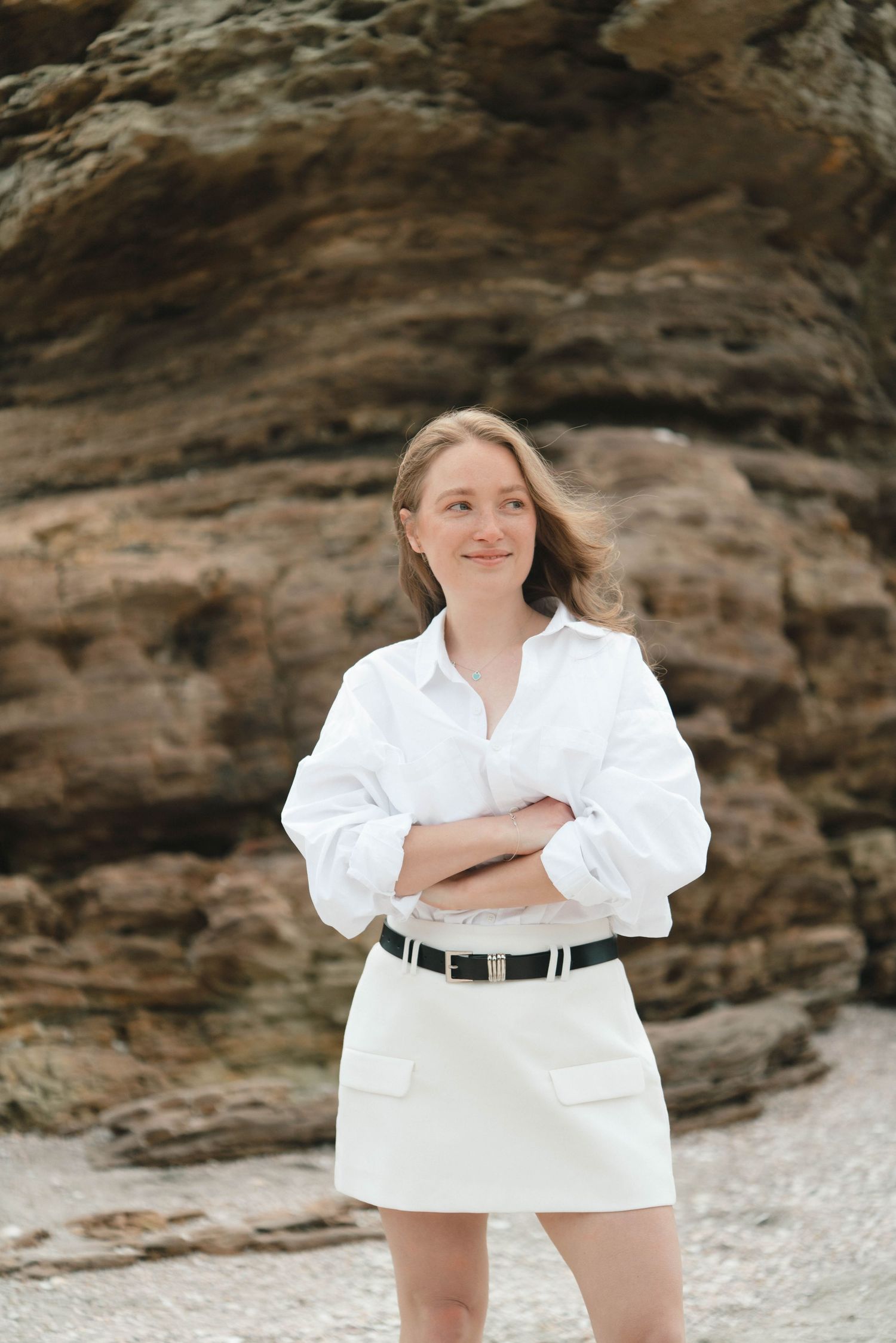Person in white dress and black belt poses confidently against rocky beach backdrop.
