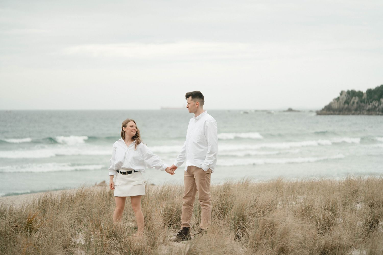 Two people in white outfits hold hands while standing on grassy dunes overlooking a beach and ocean.