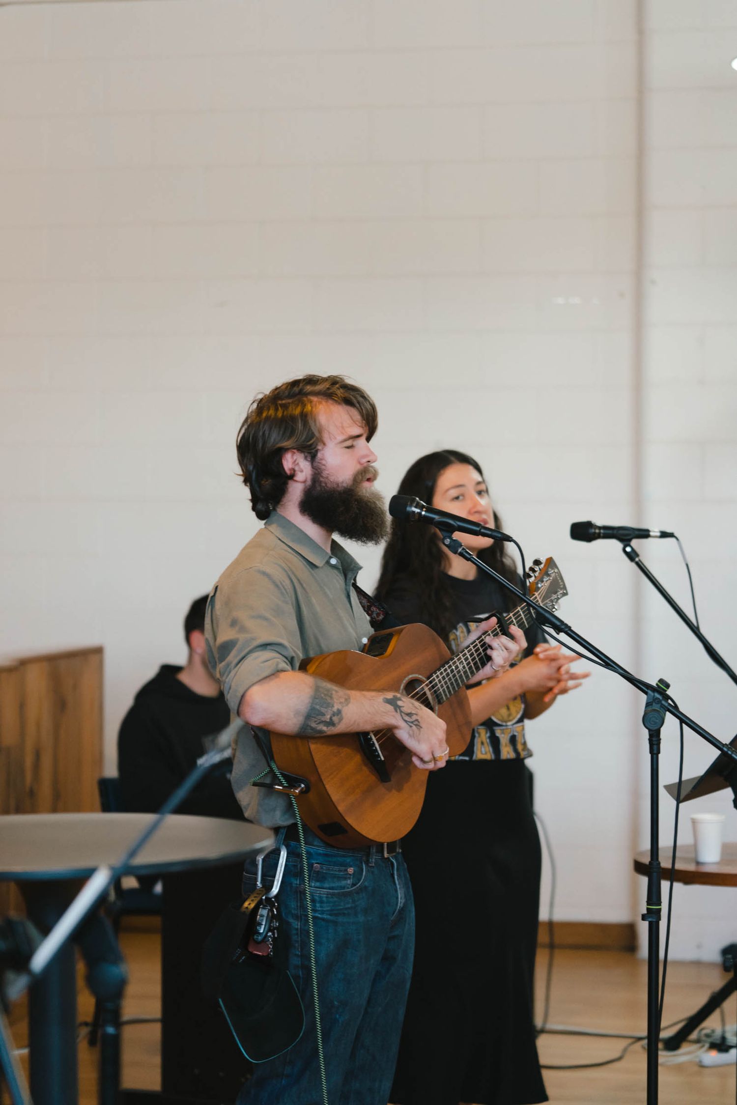 Musicians perform on stage with acoustic guitar and microphones in a minimalist indoor setting.