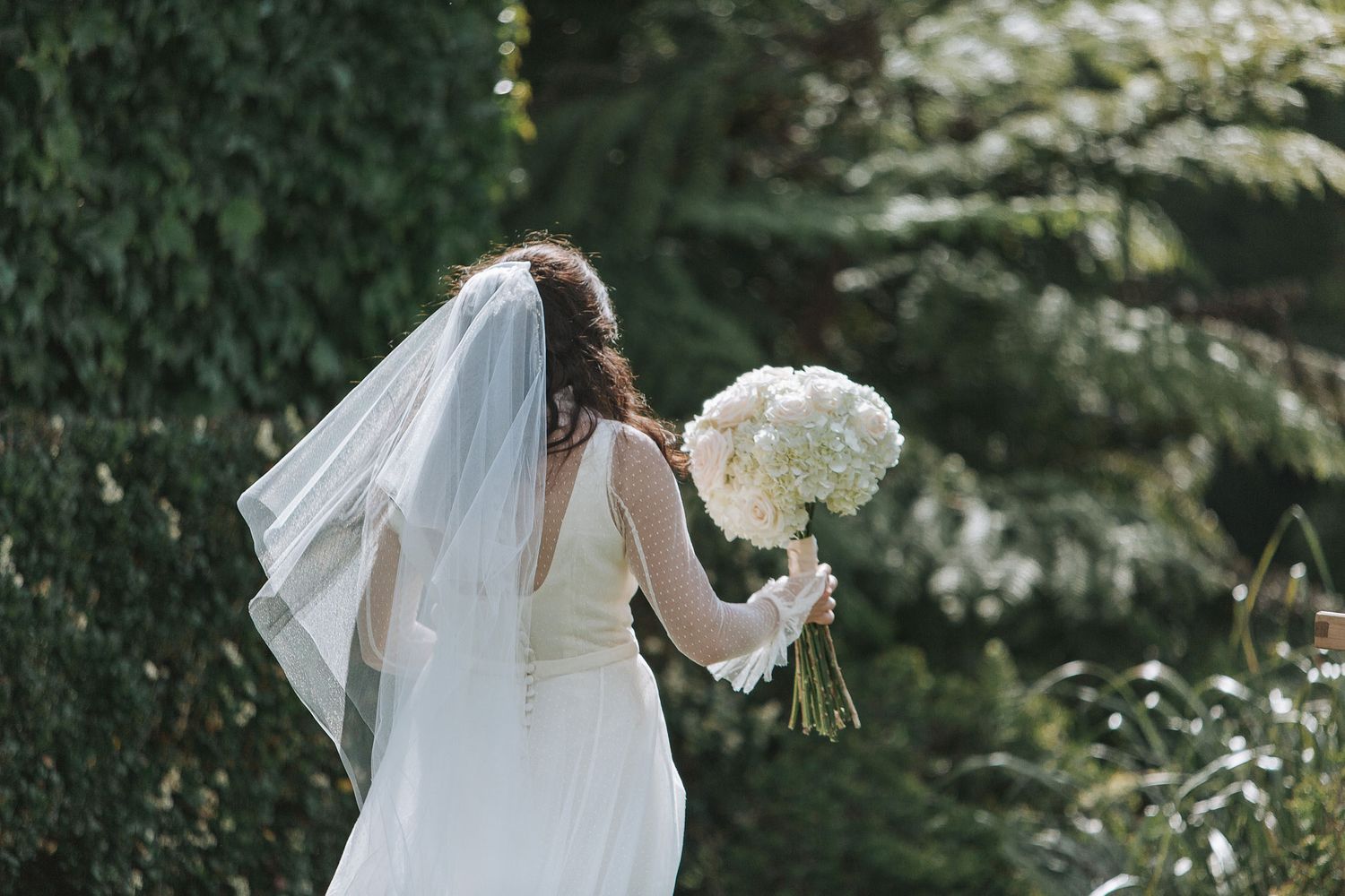A bride in a white wedding gown holds a bouquet of white flowers against a green outdoor backdrop.