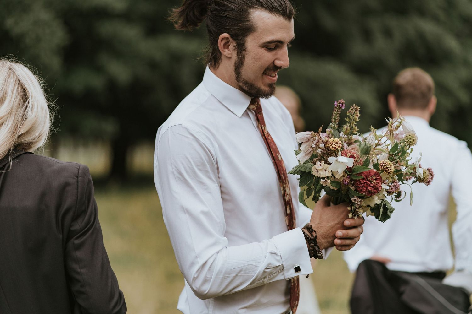 A person at a wedding celebration holds a beautiful bouquet of pink and red wildflowers in a romantic outdoor setting.