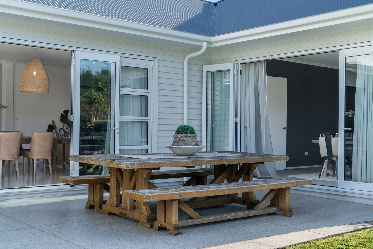 Modern outdoor dining area with wooden picnic table and benches on a concrete patio next to a grey house.