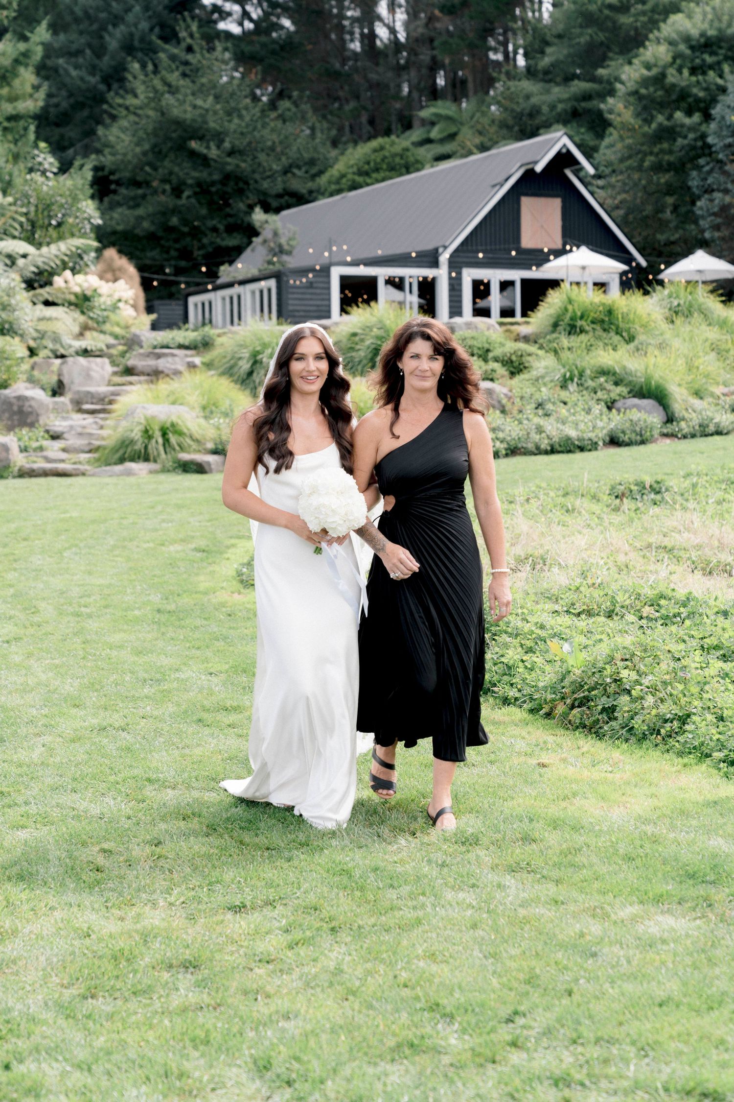 Wedding party members in black and white attire walk across a grassy lawn with a house in the background.