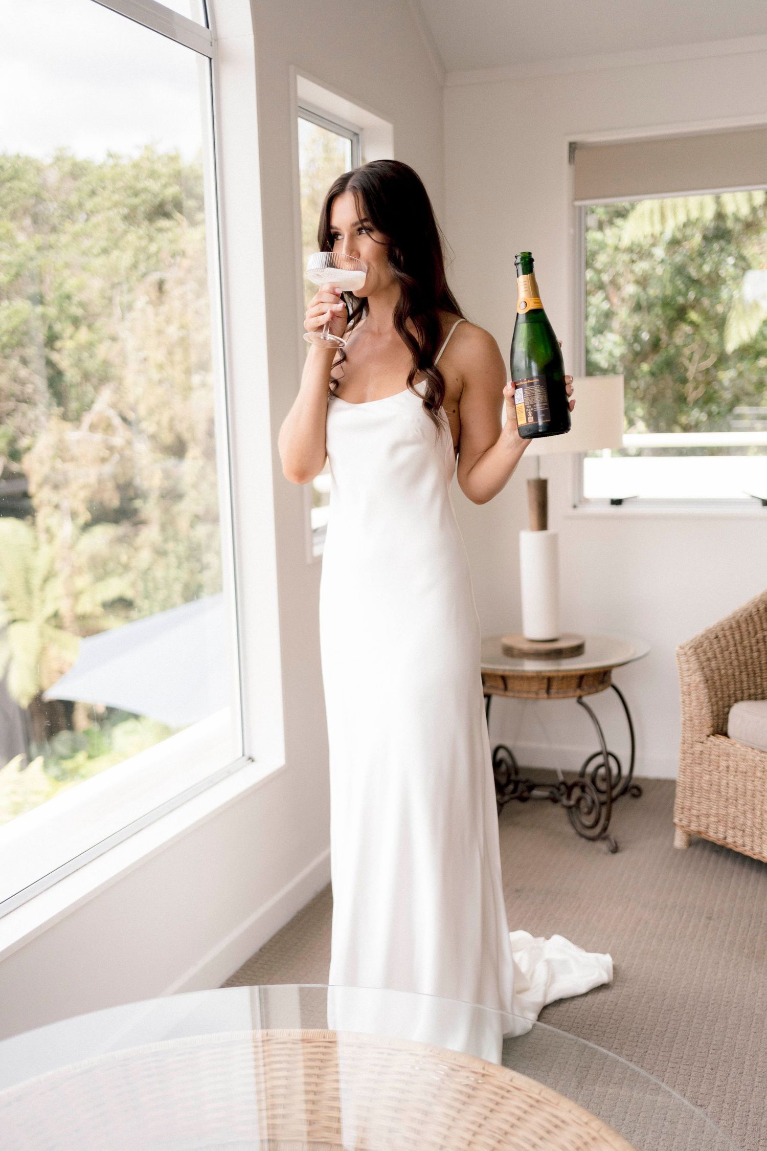 A person in a white wedding dress holds a champagne bottle while looking out a bright window.