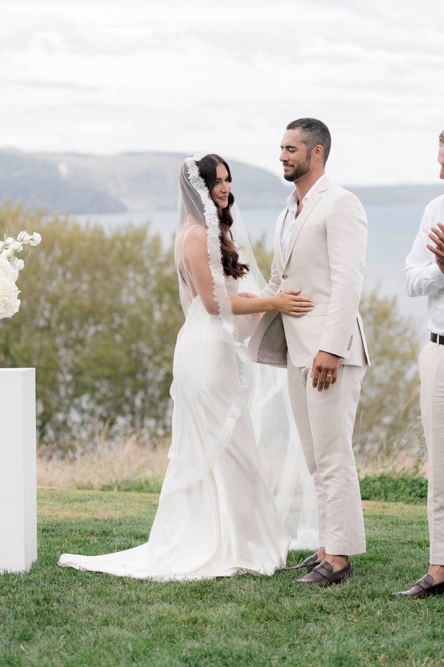 Wedding couple embraces on a grassy field with a scenic mountain backdrop.
