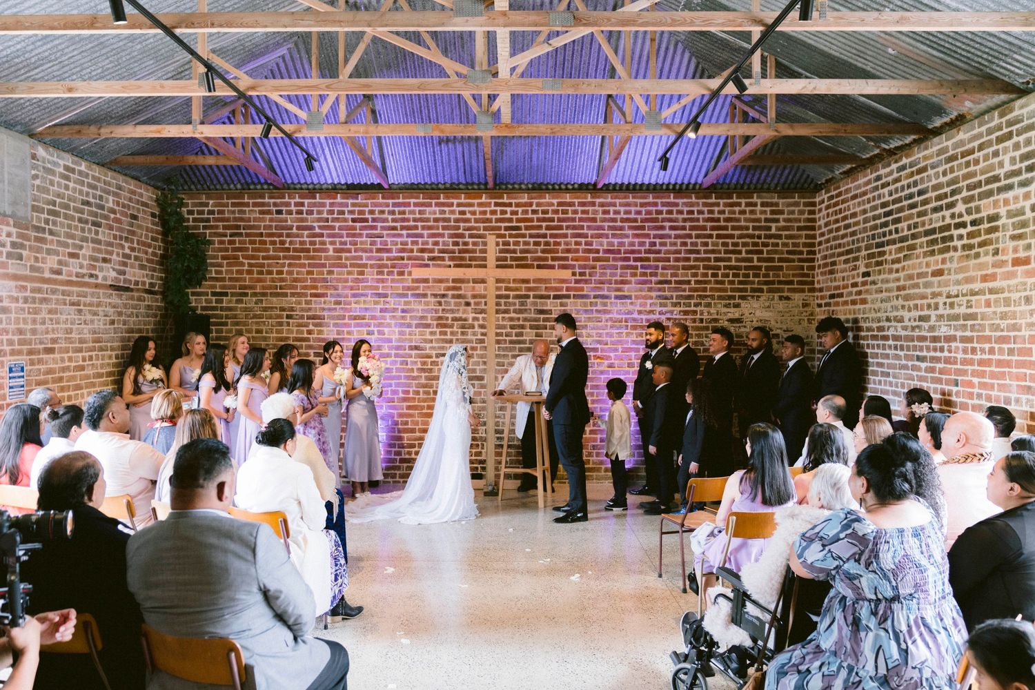 Indoor wedding ceremony in a rustic brick venue with exposed beams and purple accent lighting.