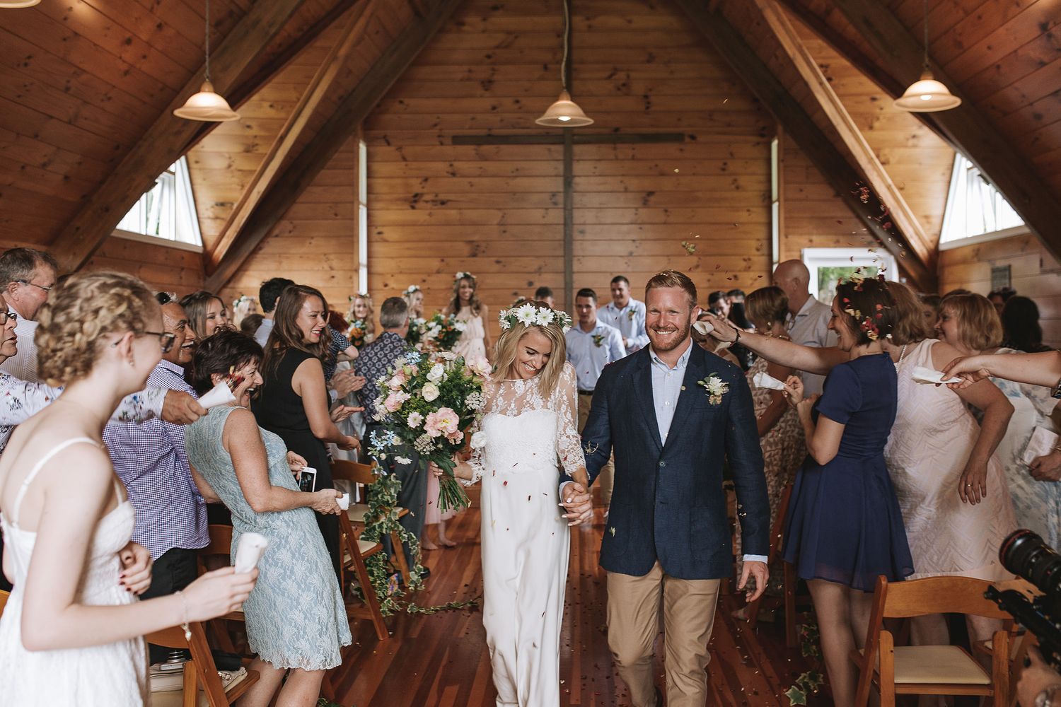 Wedding guests dance and celebrate on a rustic wood-paneled dance floor under skylights at a barn reception venue.