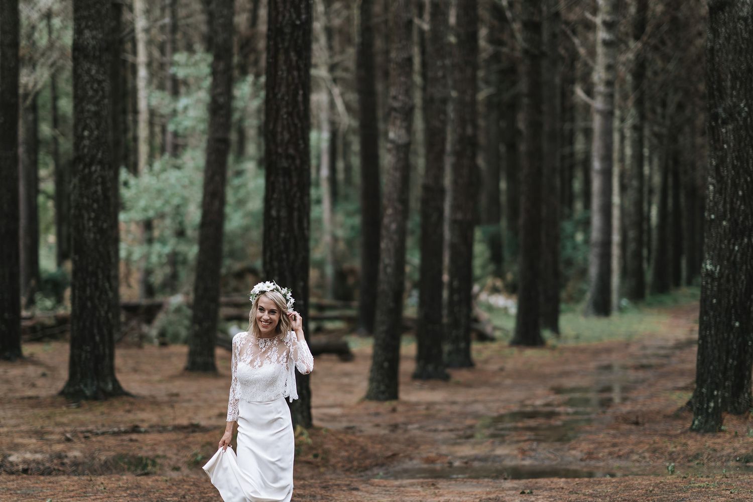 A person wearing a white dress walks through a dense pine forest with tall dark tree trunks.