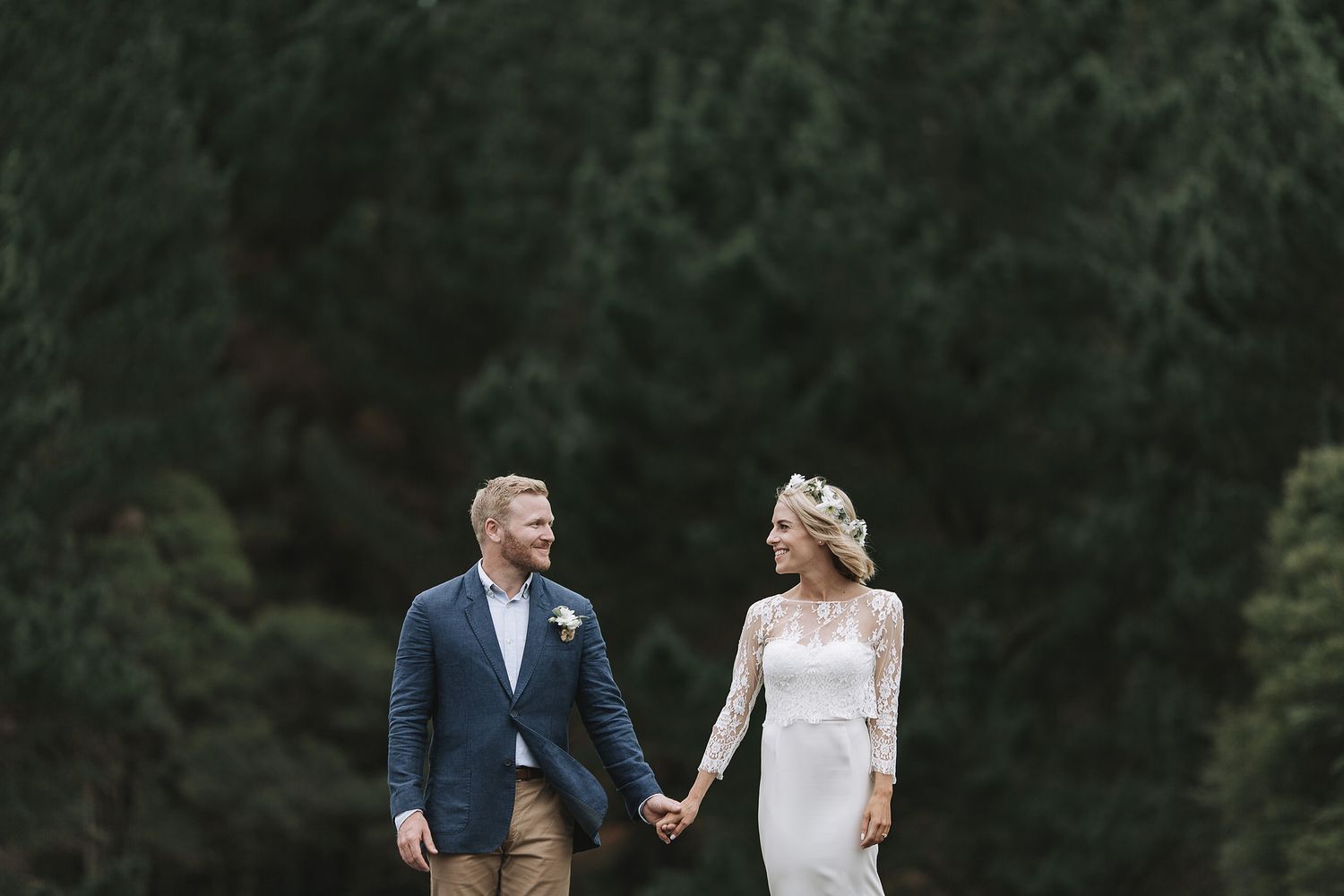 Couple holds hands while walking outdoors in romantic forest setting with bride wearing long sleeve lace gown.