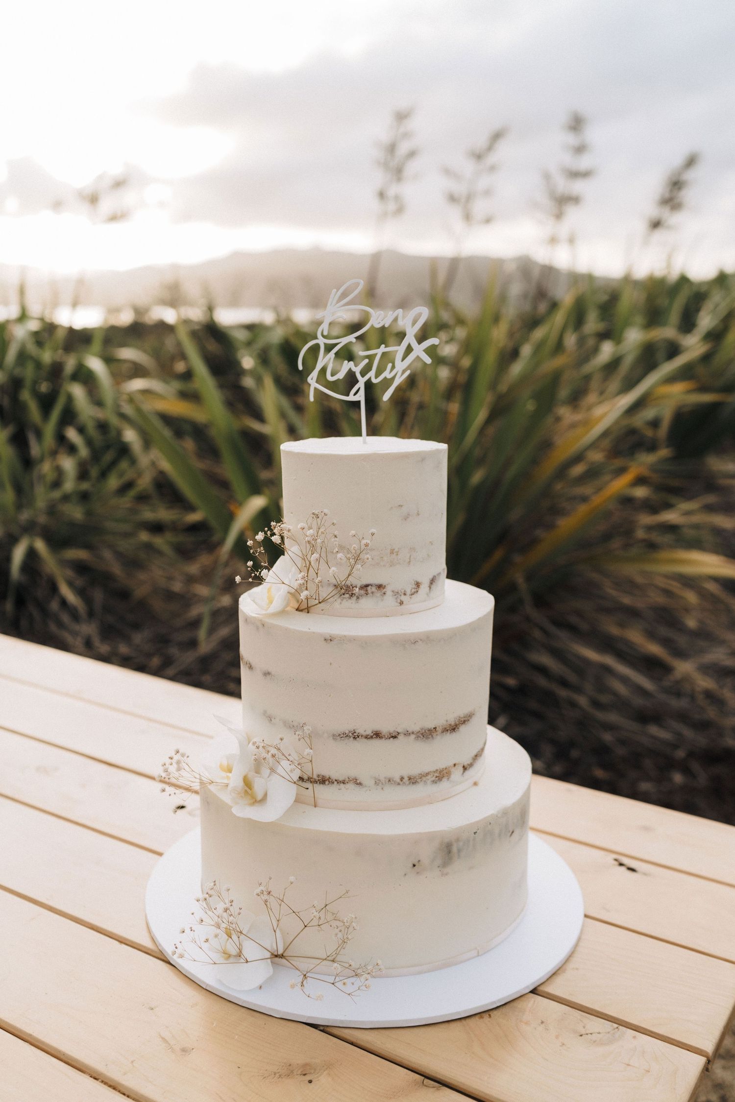Simple white two-tier wedding cake decorated with delicate dried flowers on a wooden table outdoors.