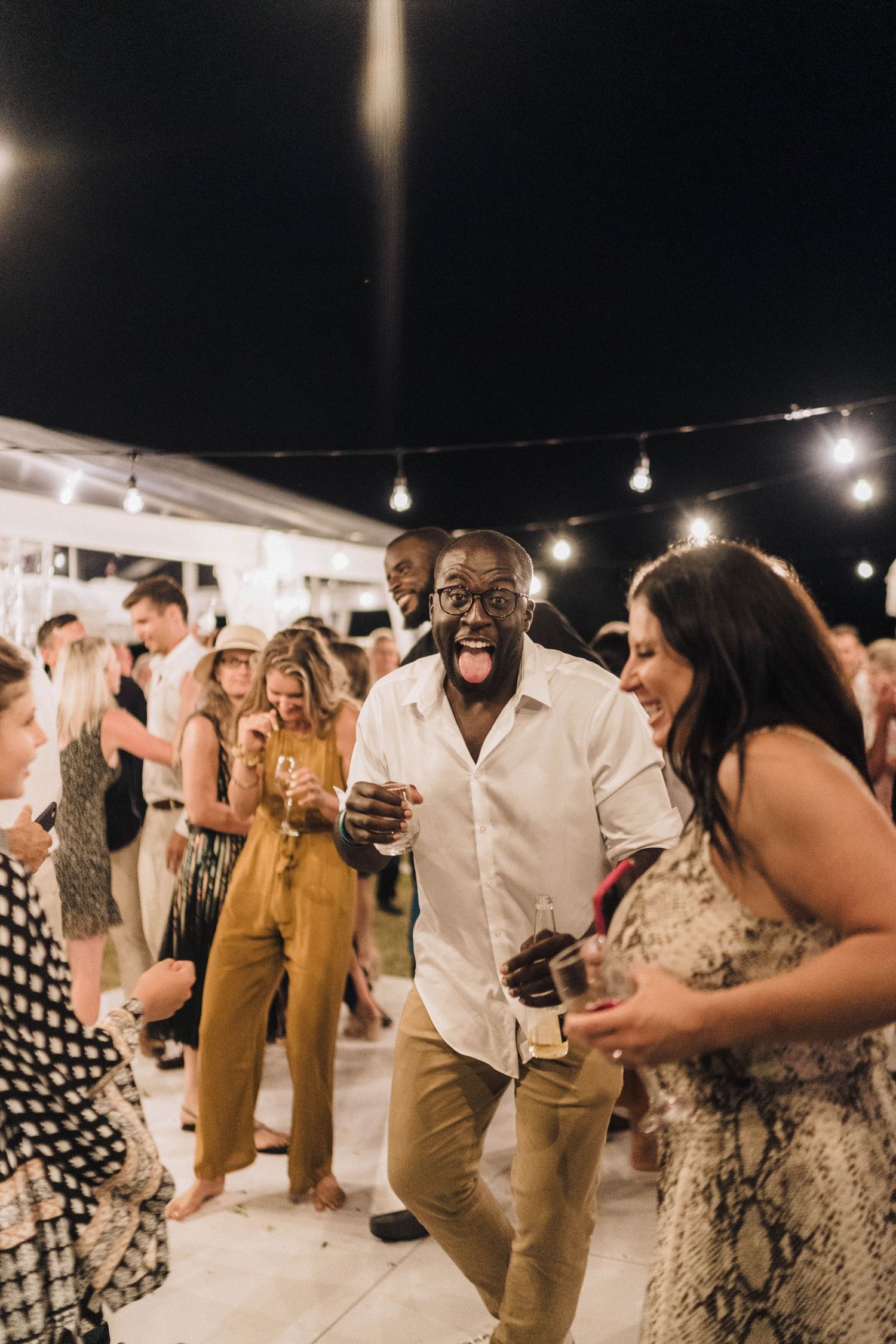 Wedding guests dance together at an outdoor reception with string lights overhead during a nighttime celebration.