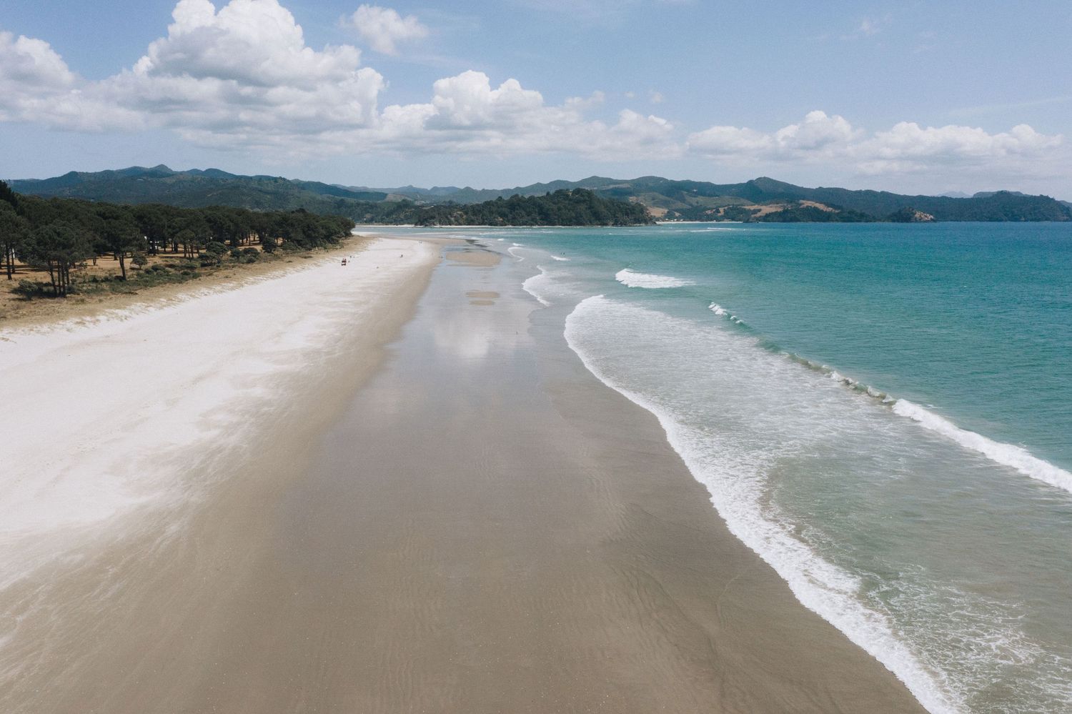 Panoramic view of a pristine curved beach with turquoise waters and lush mountains in the background under blue skies.
