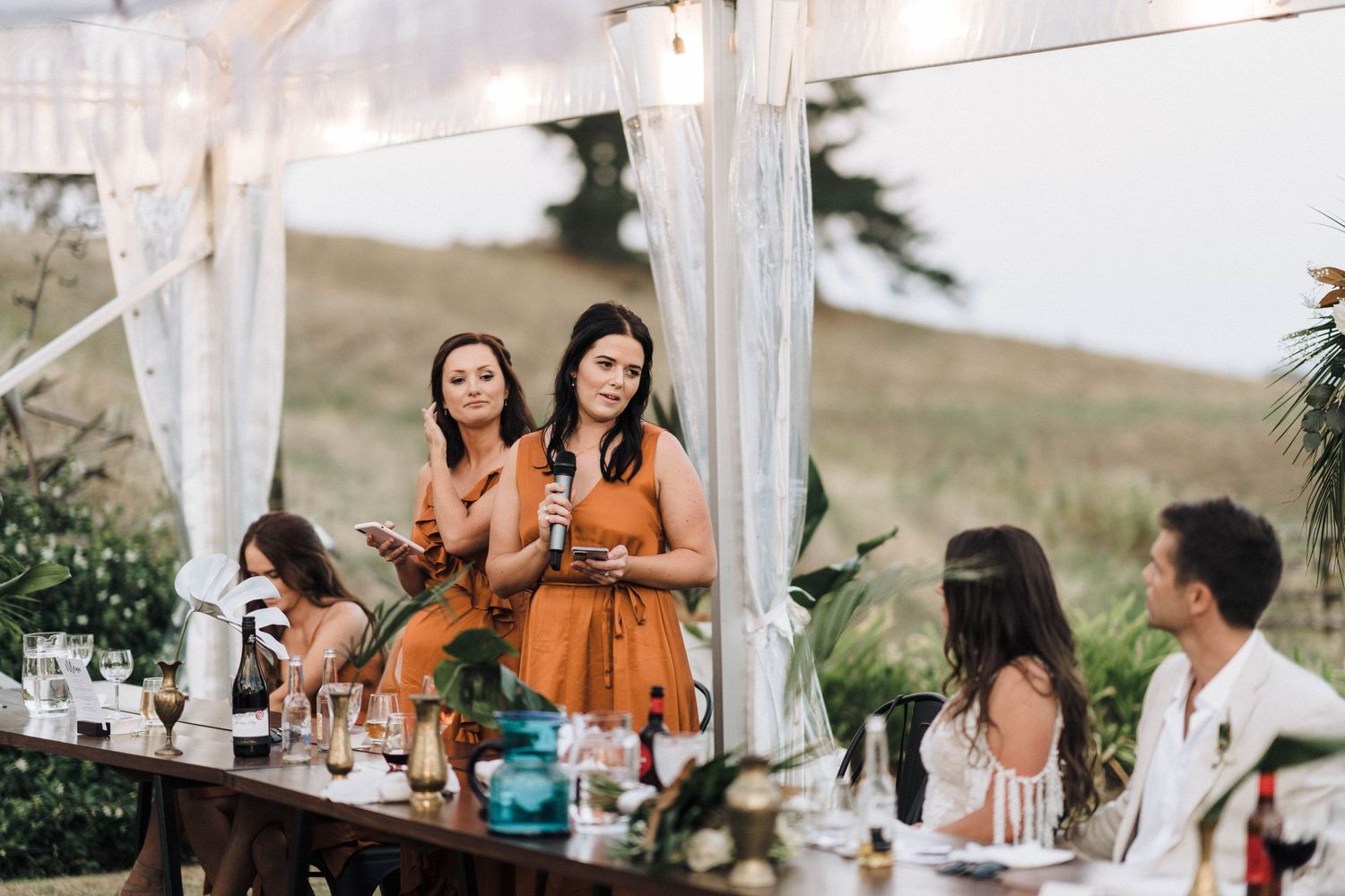 Wedding reception table with guests in orange dresses under white canopy.