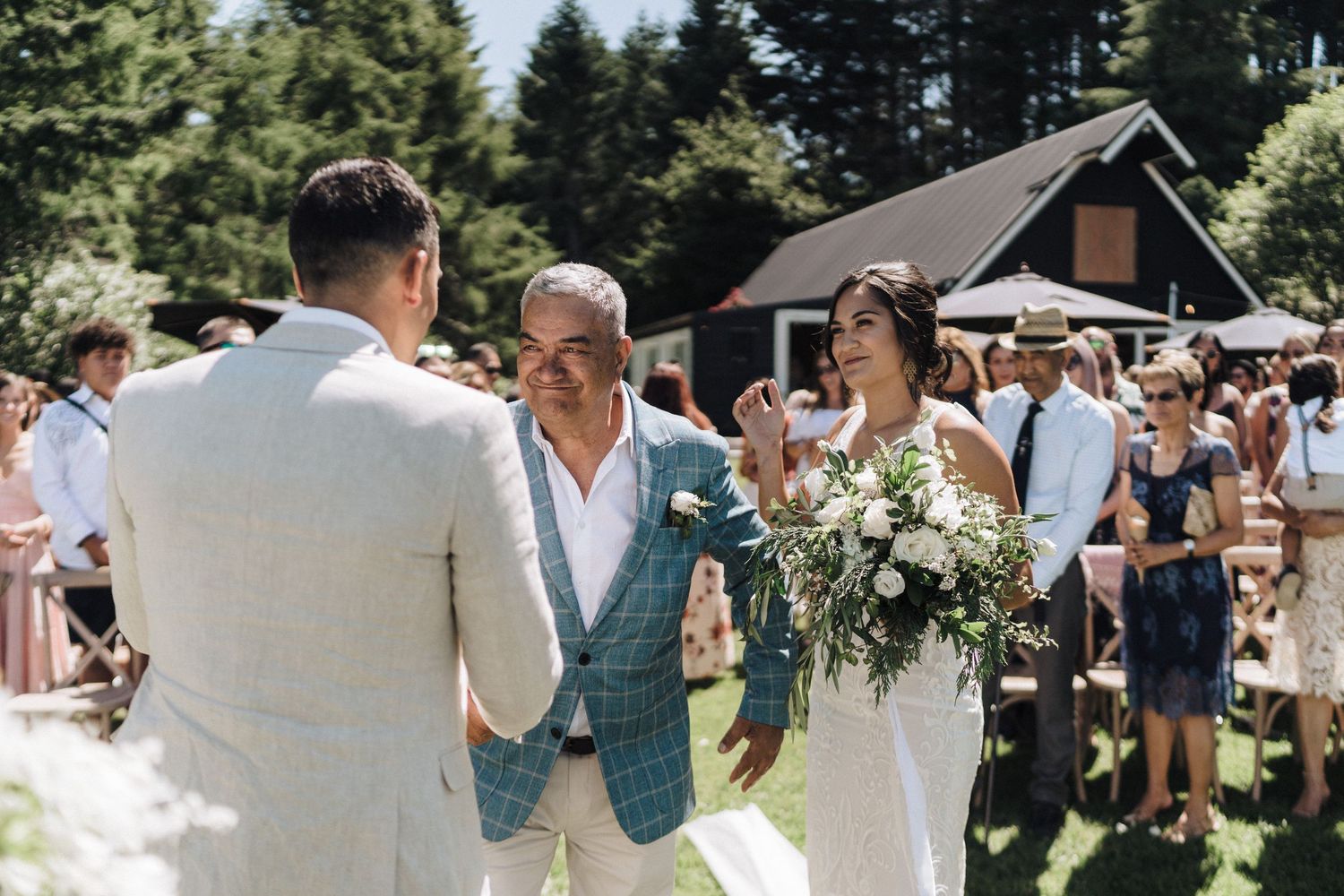 Outdoor summer wedding ceremony with guests gathered in a sunny garden setting near a cabin.