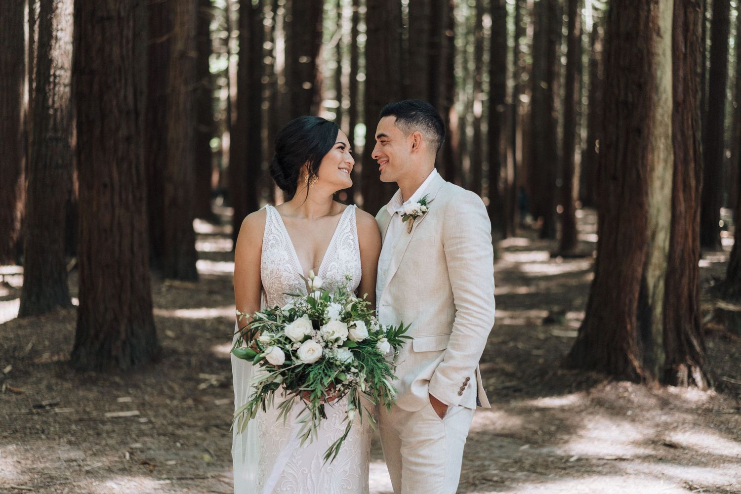 A romantic couple shares tender moments in a sunlit forest with their wedding attire and floral bouquet.
