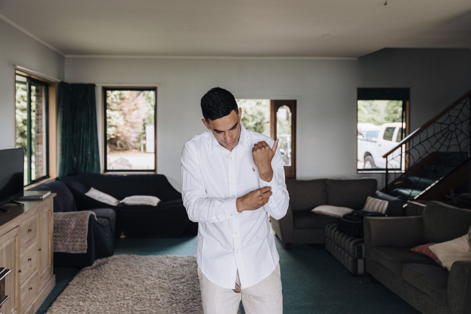 A person in all white clothing stands in a modern living room with dark furniture and natural lighting.