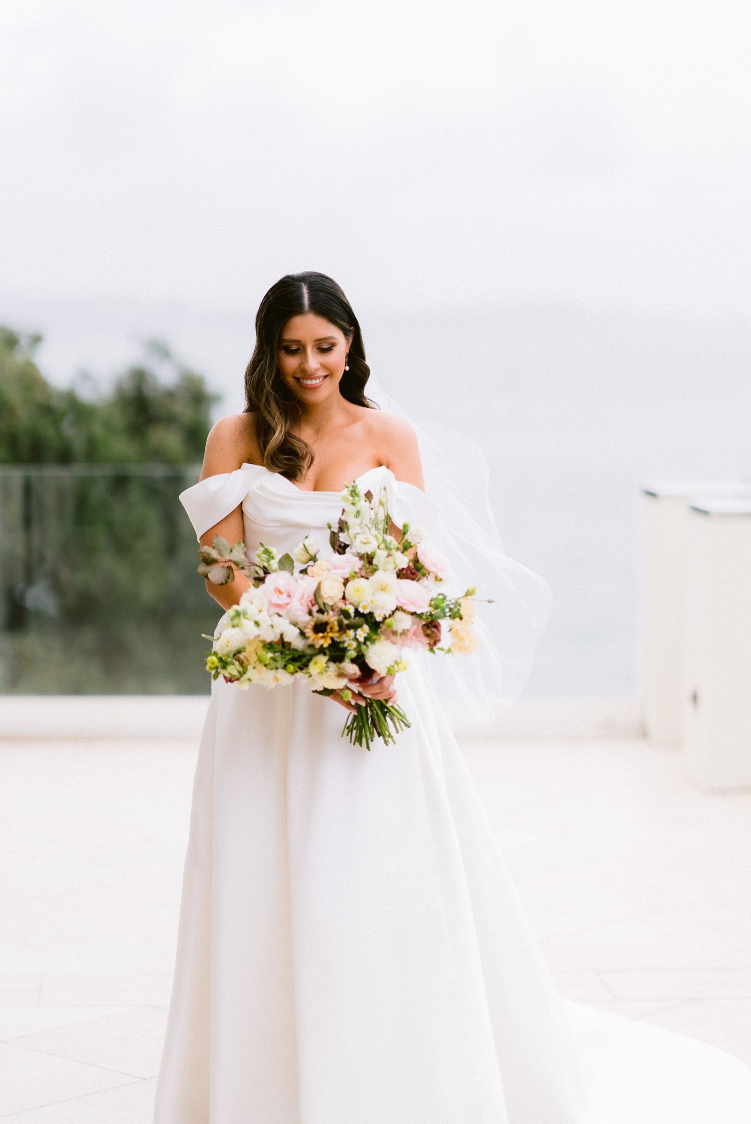 A bride in an elegant off-shoulder white gown holds a beautiful bouquet of white and pink flowers.
