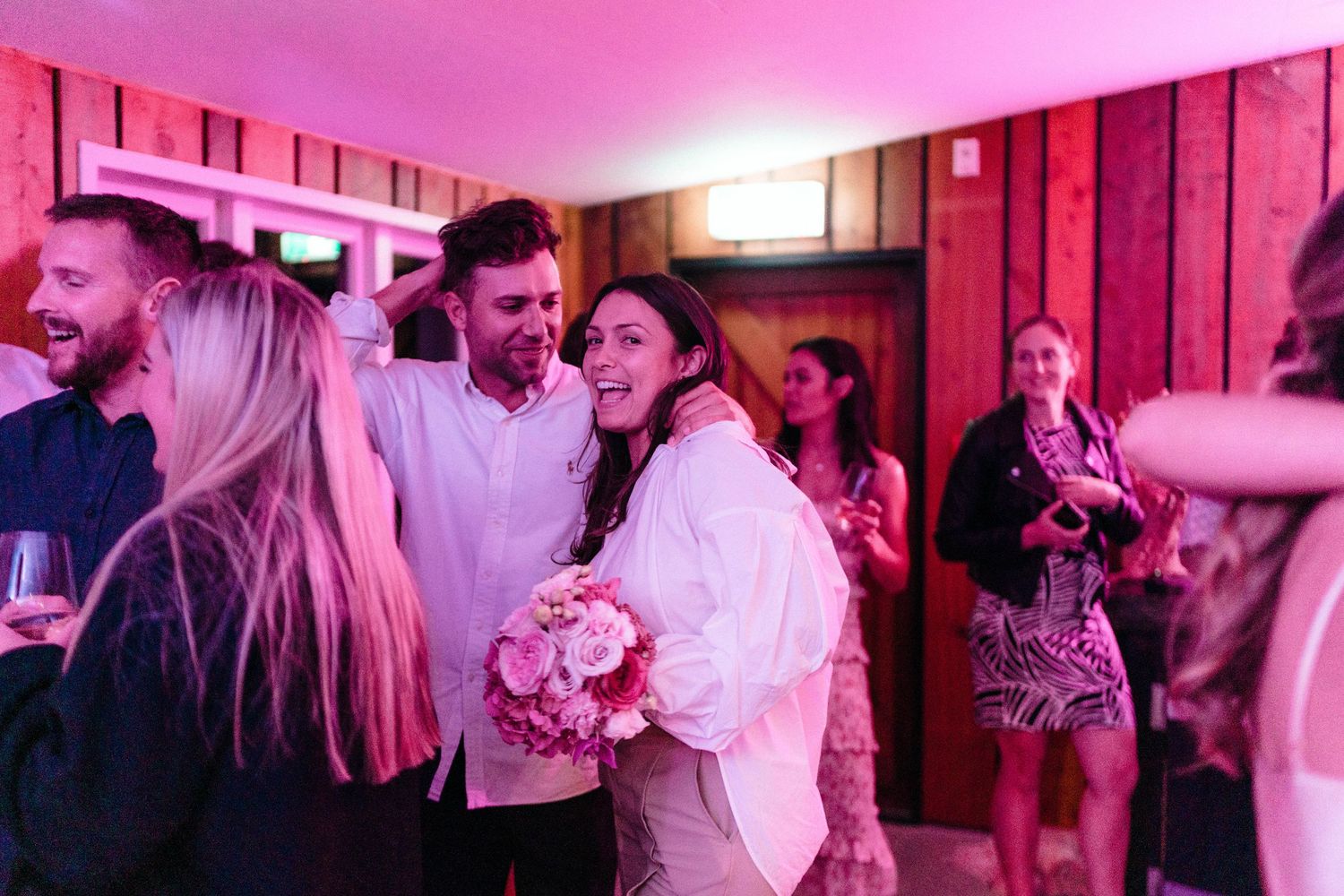 Wedding guests celebrate and dance together under pink mood lighting in a rustic wood-paneled reception space.