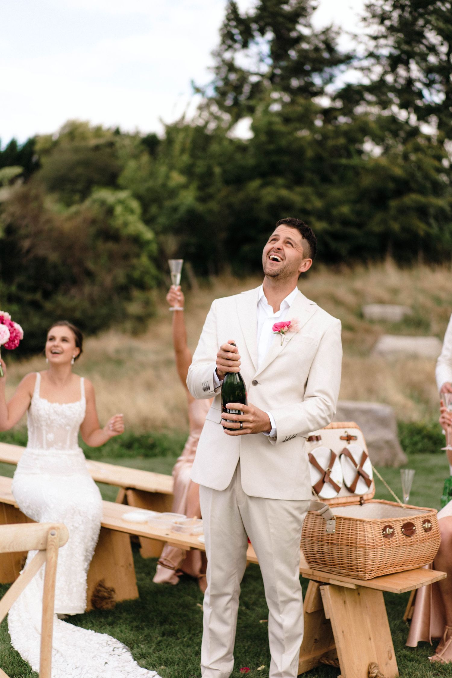 Couple enjoying drinks and laughter at outdoor wedding reception with rustic wooden benches.