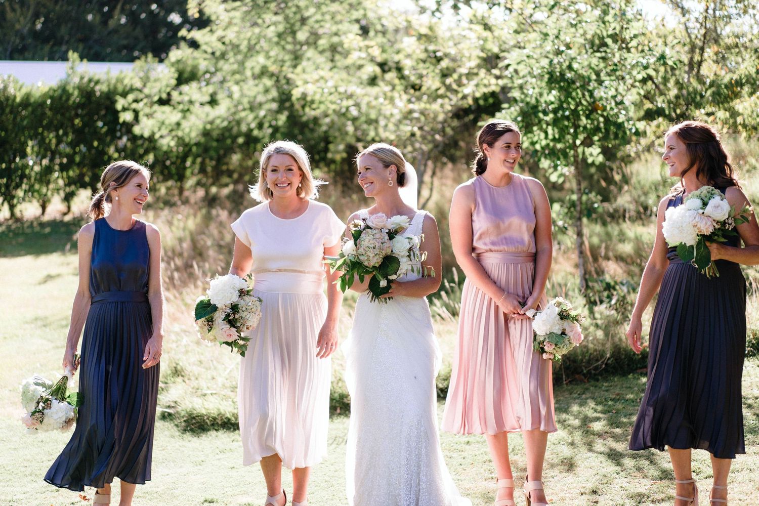 Bridal party in navy and blush pink dresses poses together during an outdoor summer celebration.