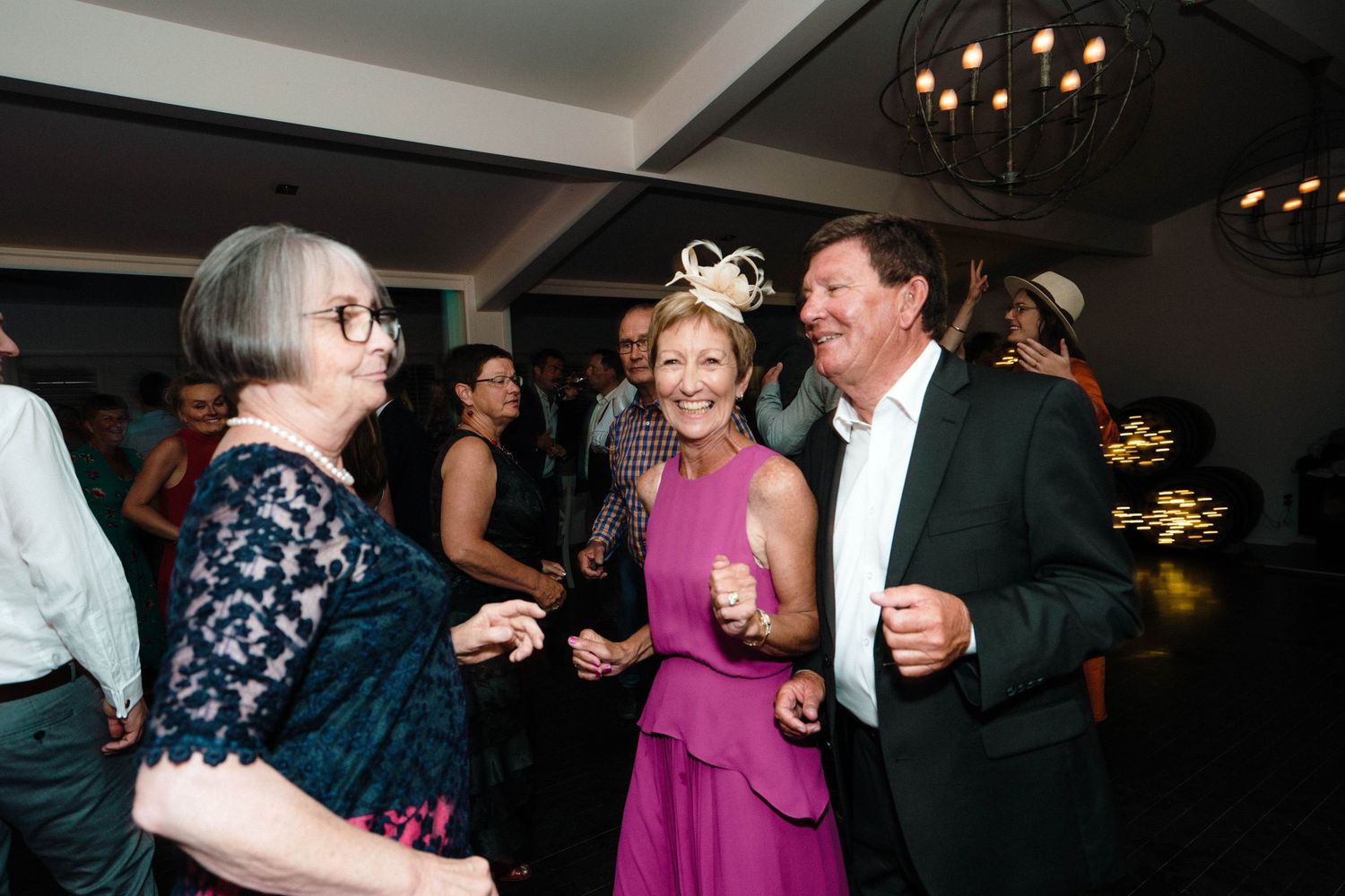 A group of guests enjoy conversation and laughter at an indoor reception with chandelier lighting overhead.