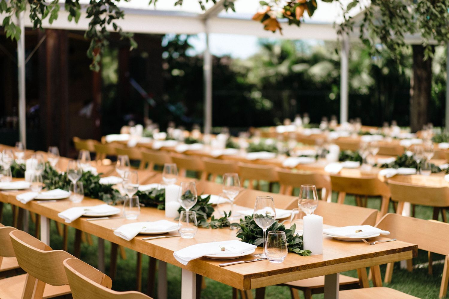 Wooden dining tables with place settings arranged outdoors under natural lighting for an elegant event setup.