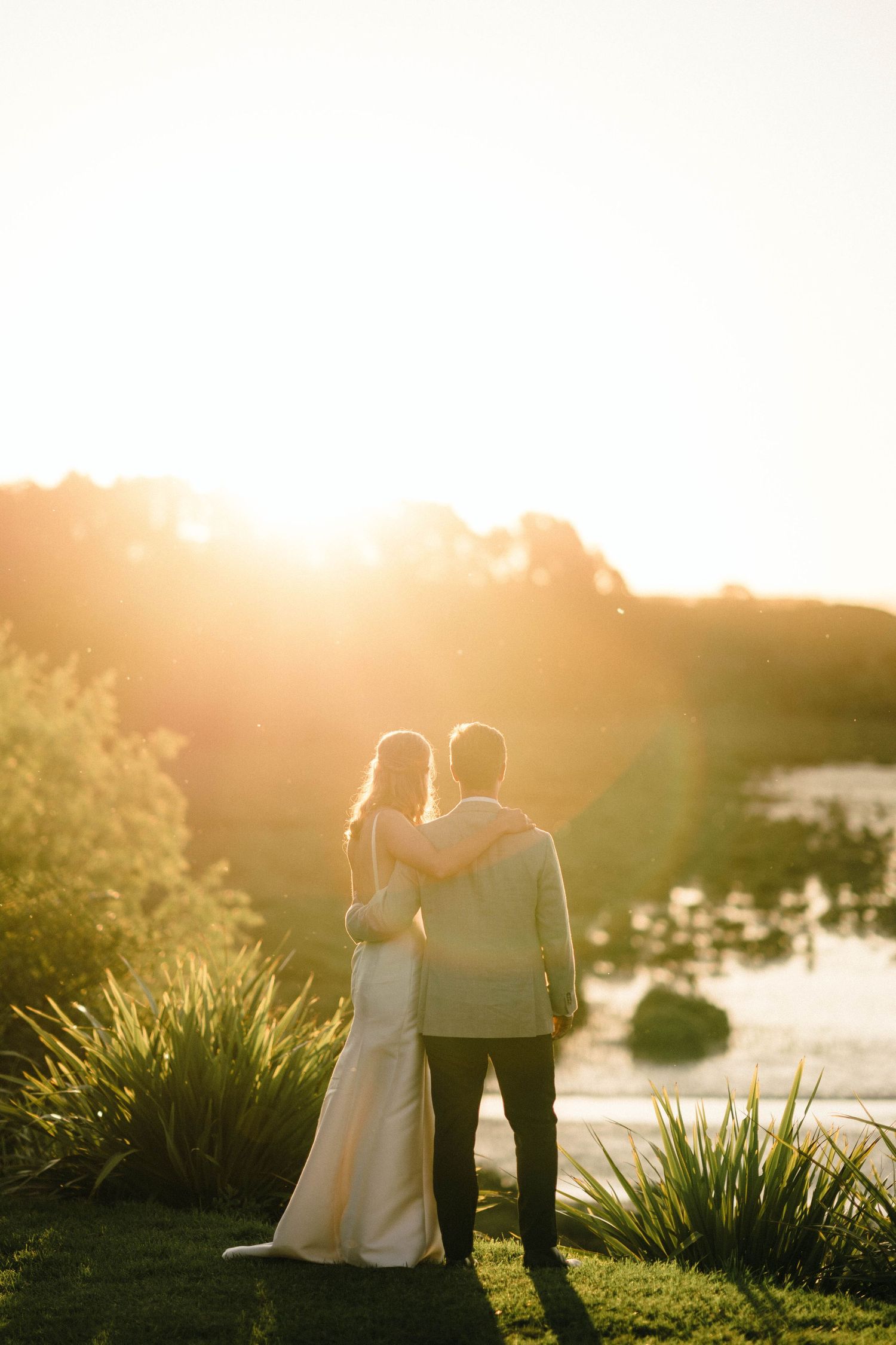 A couple embraces while watching the sunset over a pond, backlit by golden hour light among decorative grasses.
