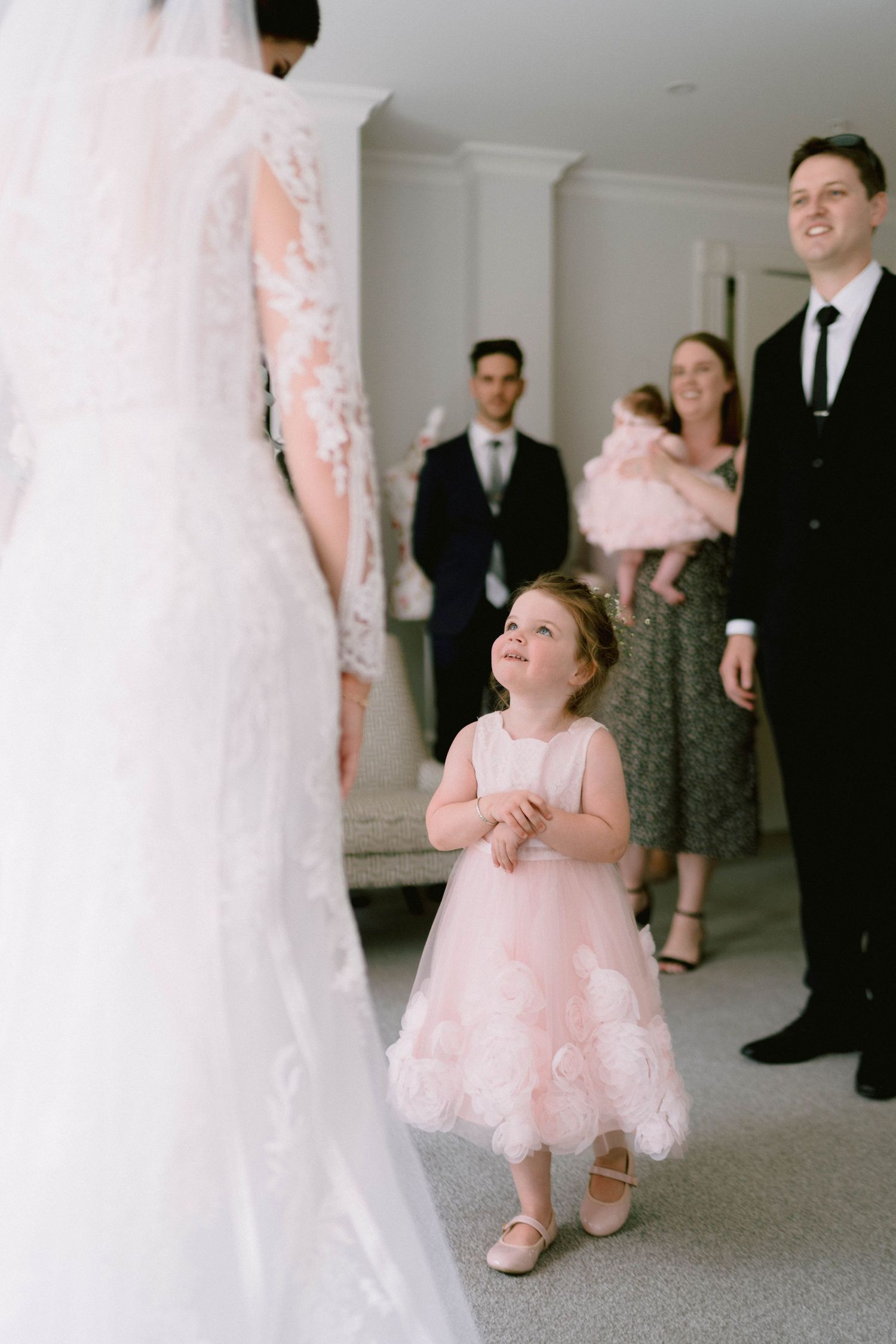 A young flower girl in pink dress looks up in awe at a bride in her wedding gown.