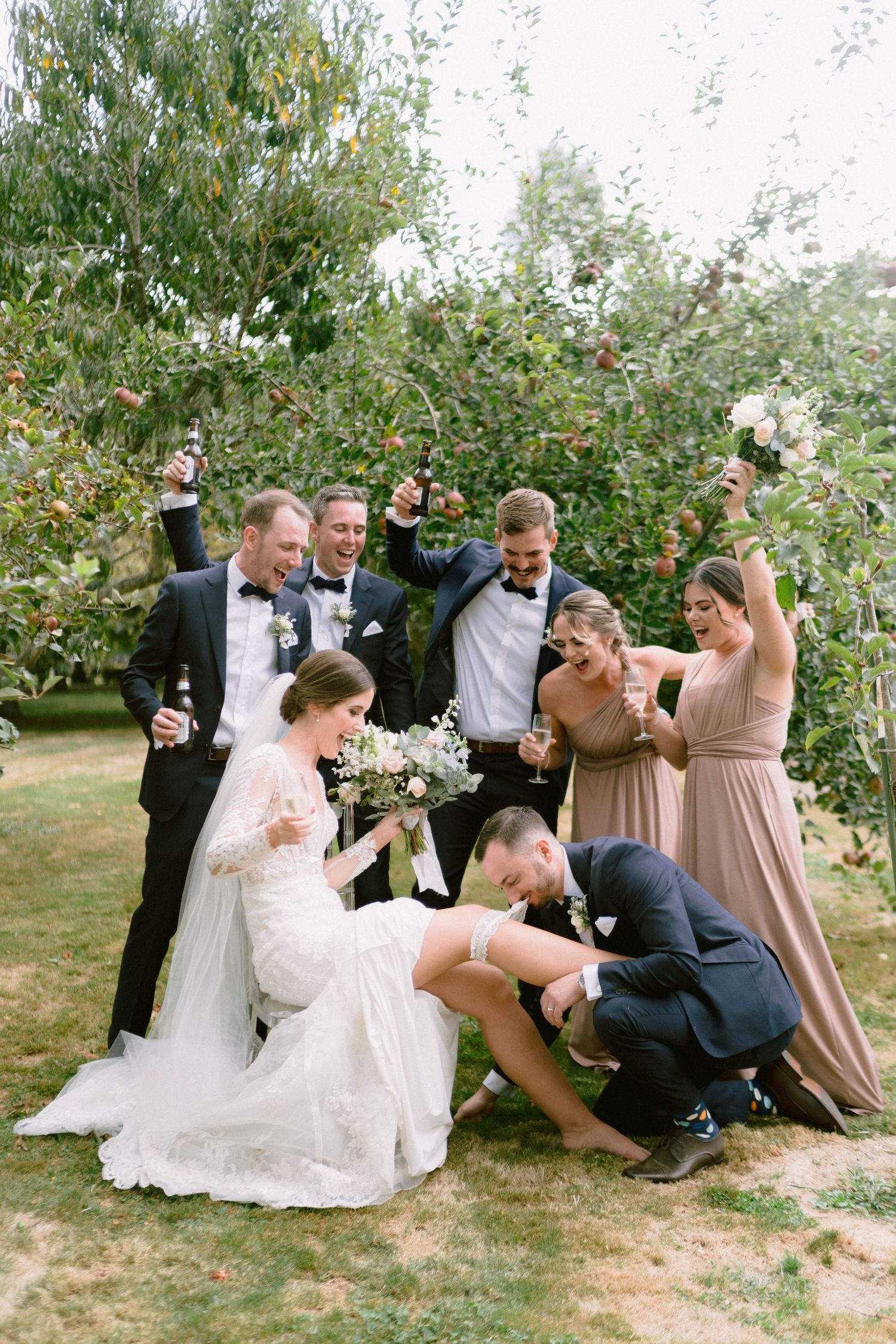 Wedding party members sitting and laughing together in an apple orchard.