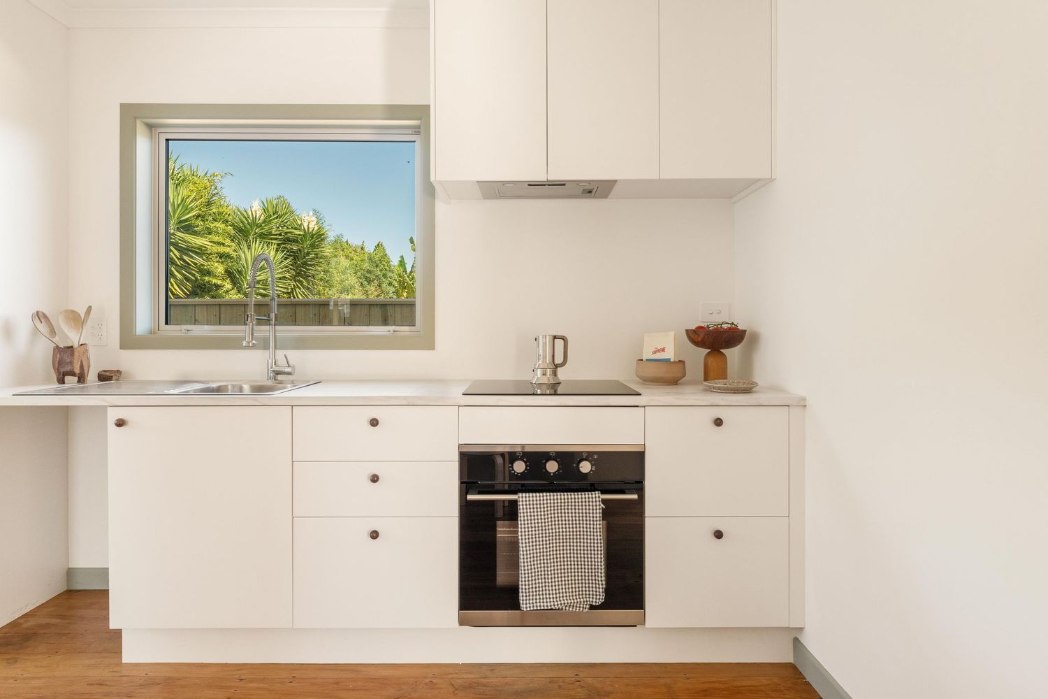 Minimalist white kitchen featuring built-in appliances and window overlooking green trees.