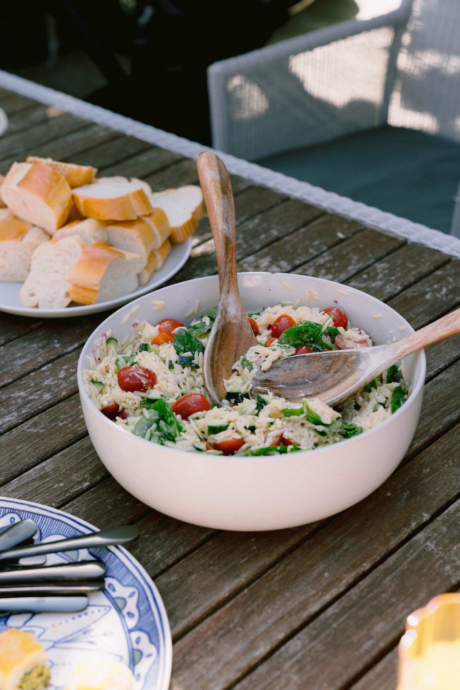 Fresh pasta salad with cherry tomatoes and broccoli in pink bowl alongside bread plate on wooden outdoor table.