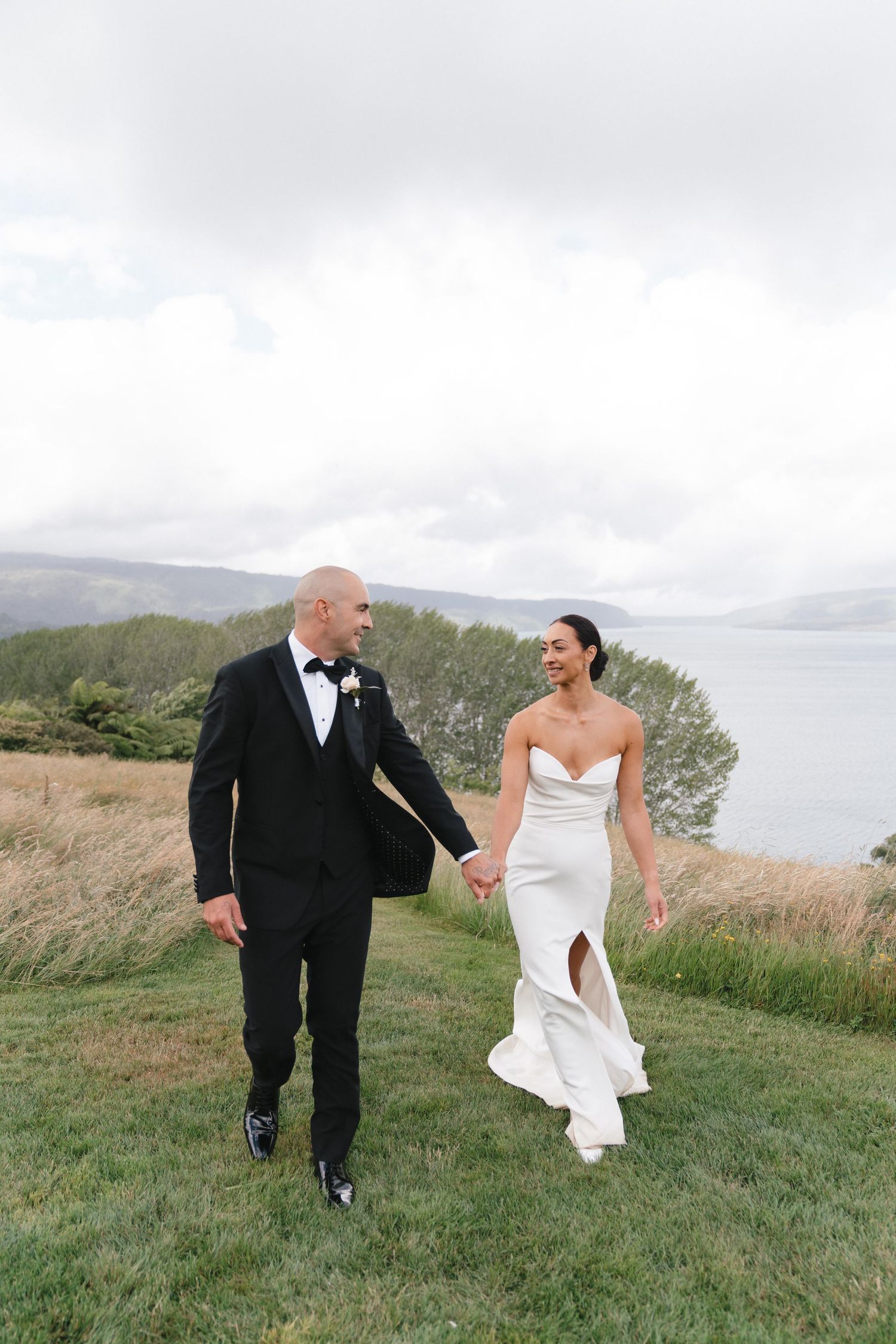Bride and groom walk and laugh together on grassy hillside during their wedding day.