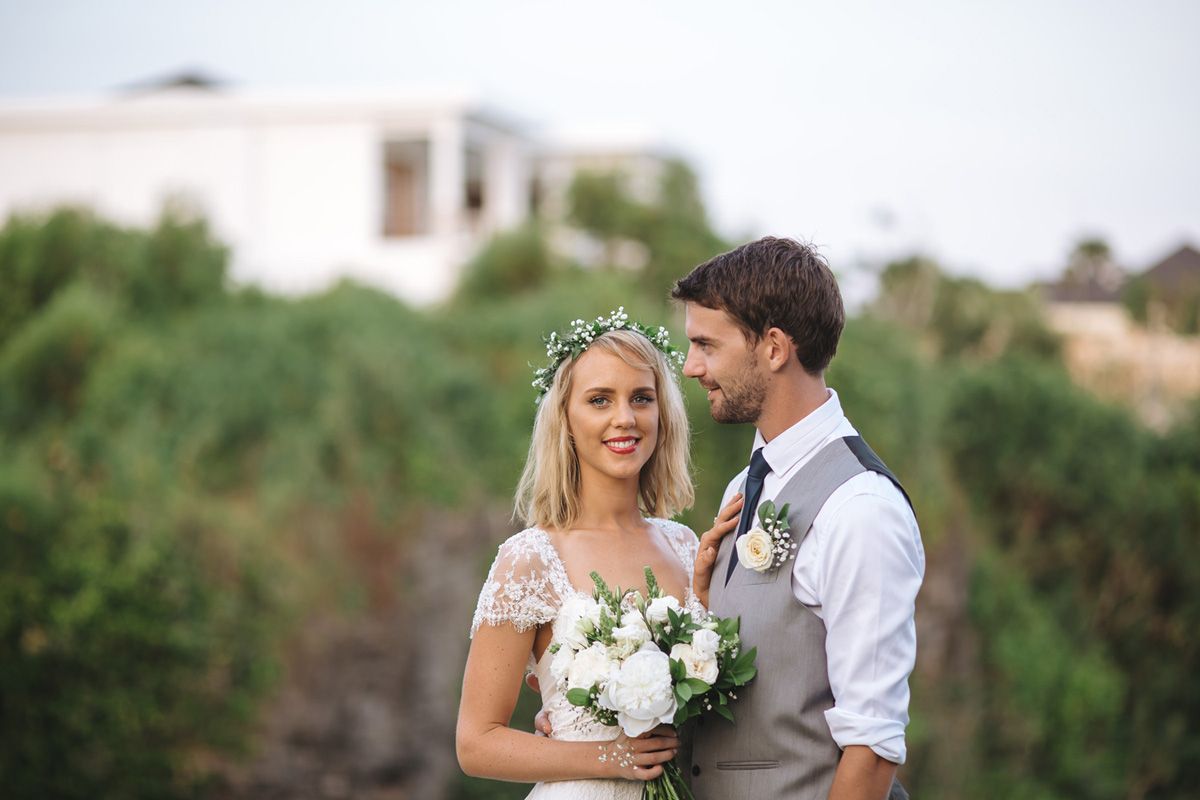 A couple in wedding attire poses together outdoors with greenery and buildings in background.