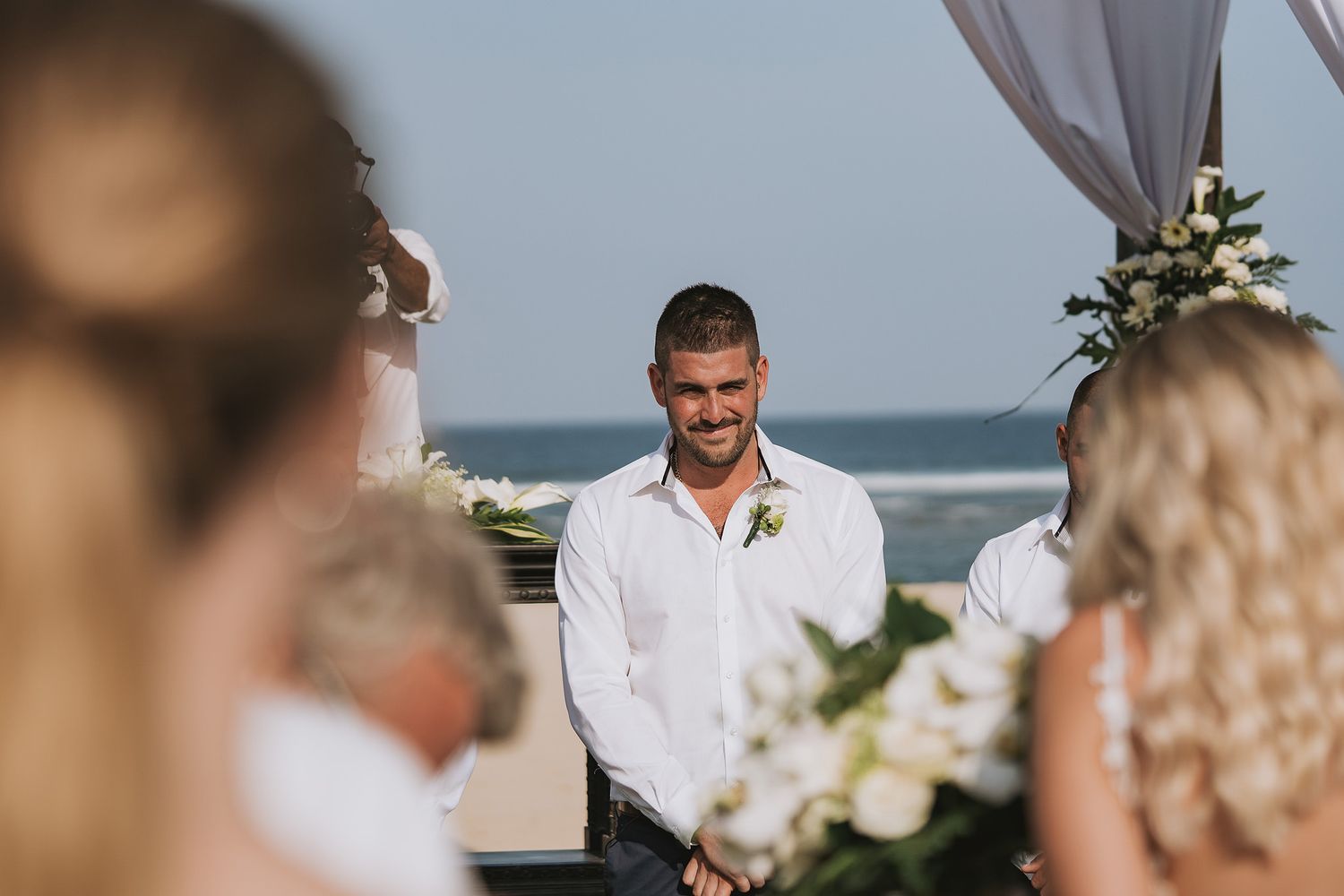 A wedding ceremony by the ocean with a white-clad groom standing under a floral-decorated arch.
