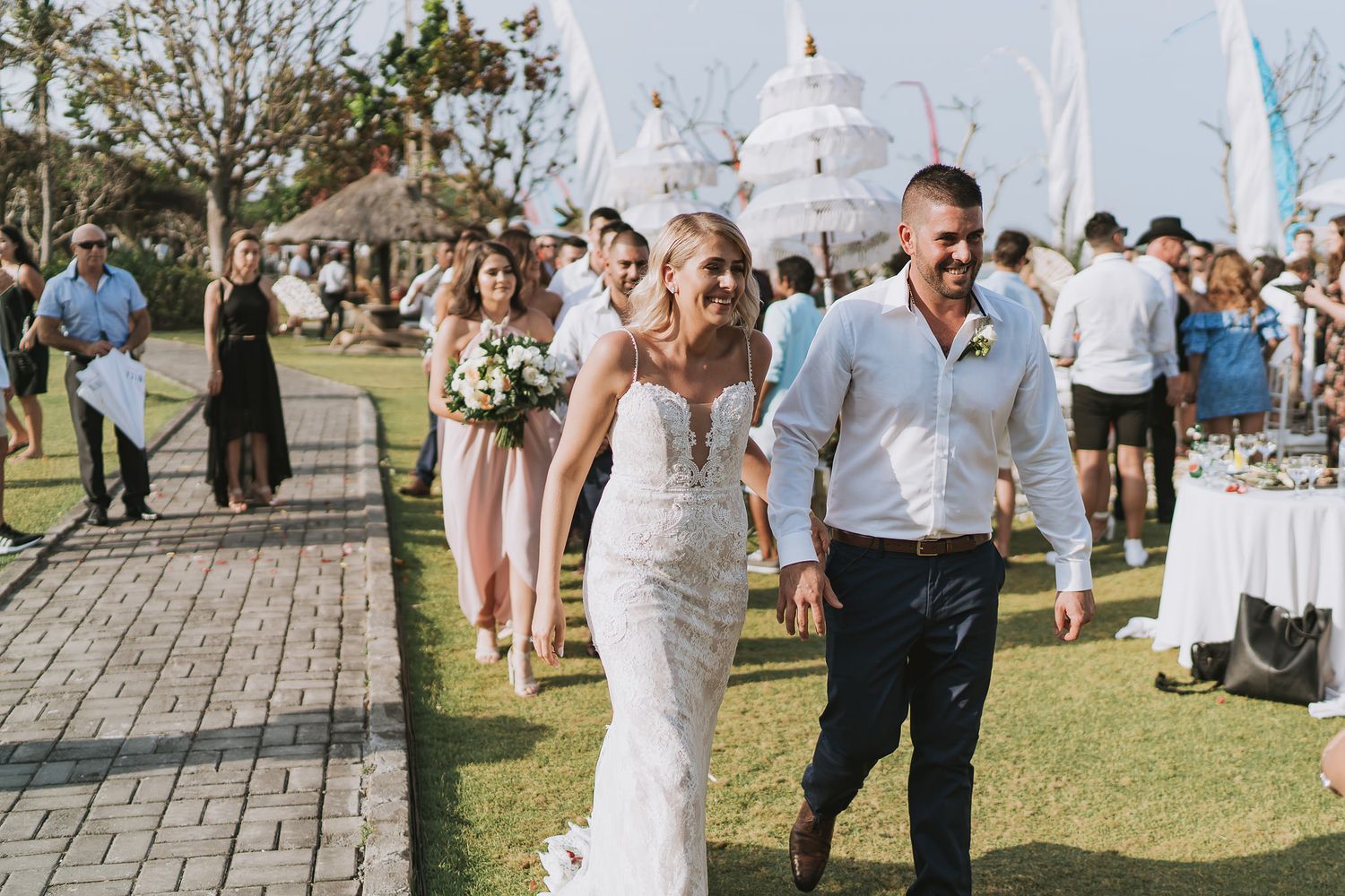 Newlyweds walking together at outdoor reception with white decor and palm trees.