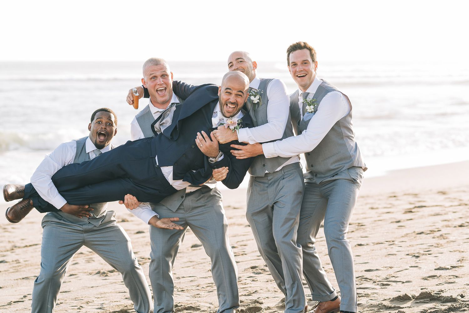 Groomsmen playfully carrying and posing on the beach in gray suits.