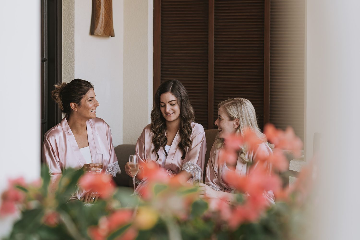 Three people in robes share a joyful conversation on a porch with coral-colored flowers in the foreground.