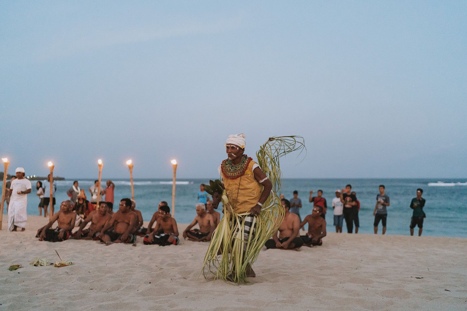 Traditional beach ceremony at sunset with performers and spectators gathered on the sand with glowing torches.