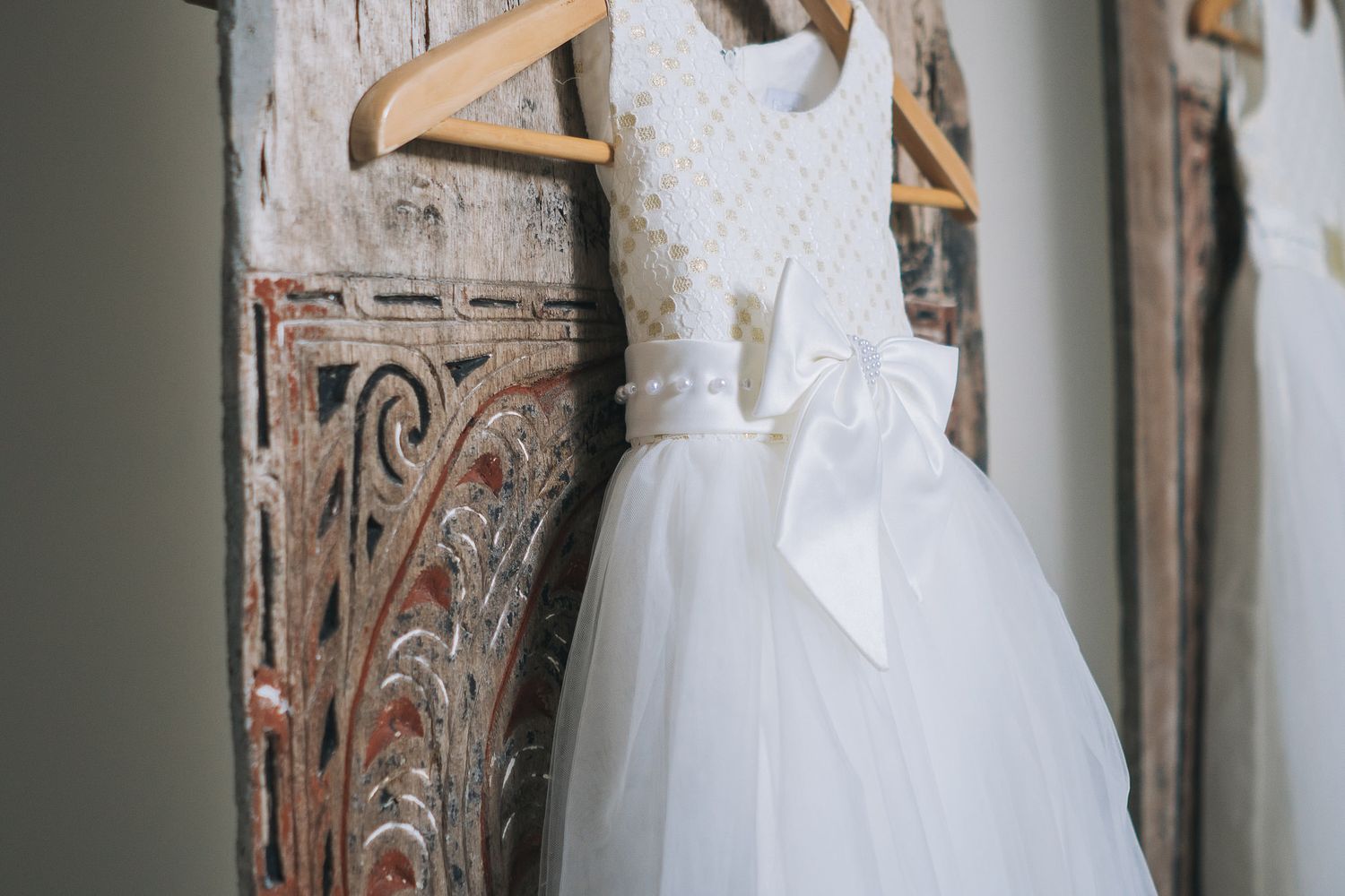 White wedding dress hanging on wooden hangers against rustic wall.