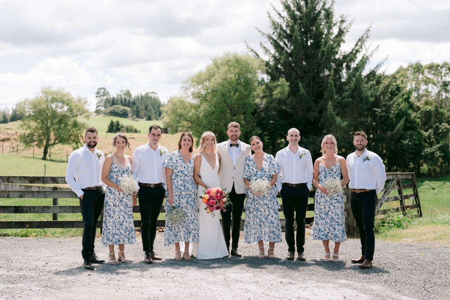 A wedding party poses together on a gravel path with bridesmaids in floral dresses and wooden fences in the background.