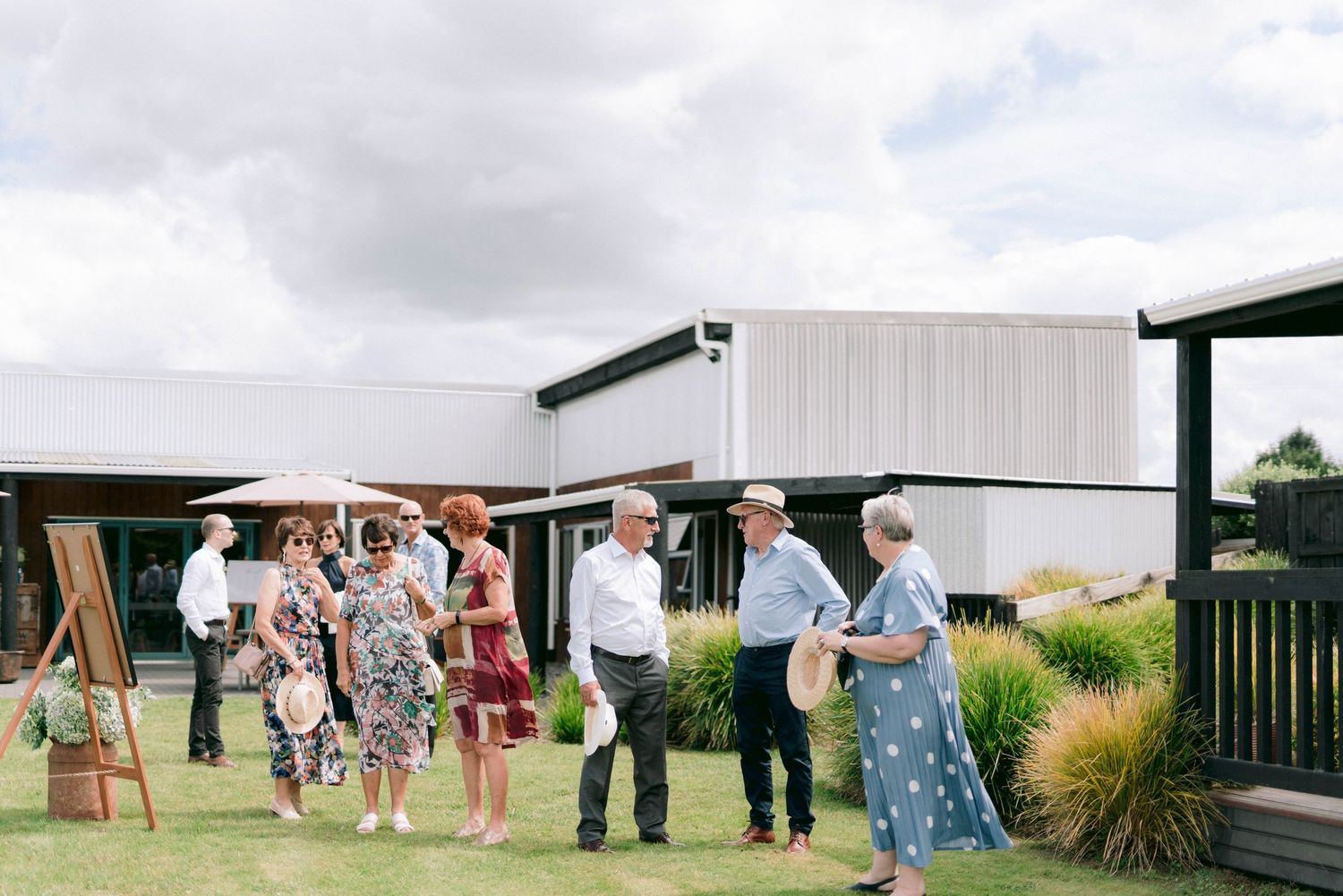 Wedding guests gather outdoors in front of white rural buildings during a sunny summer celebration.