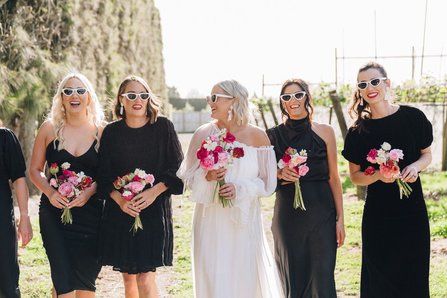 A bridal party wearing black dresses and sunglasses pose together holding pink floral bouquets.