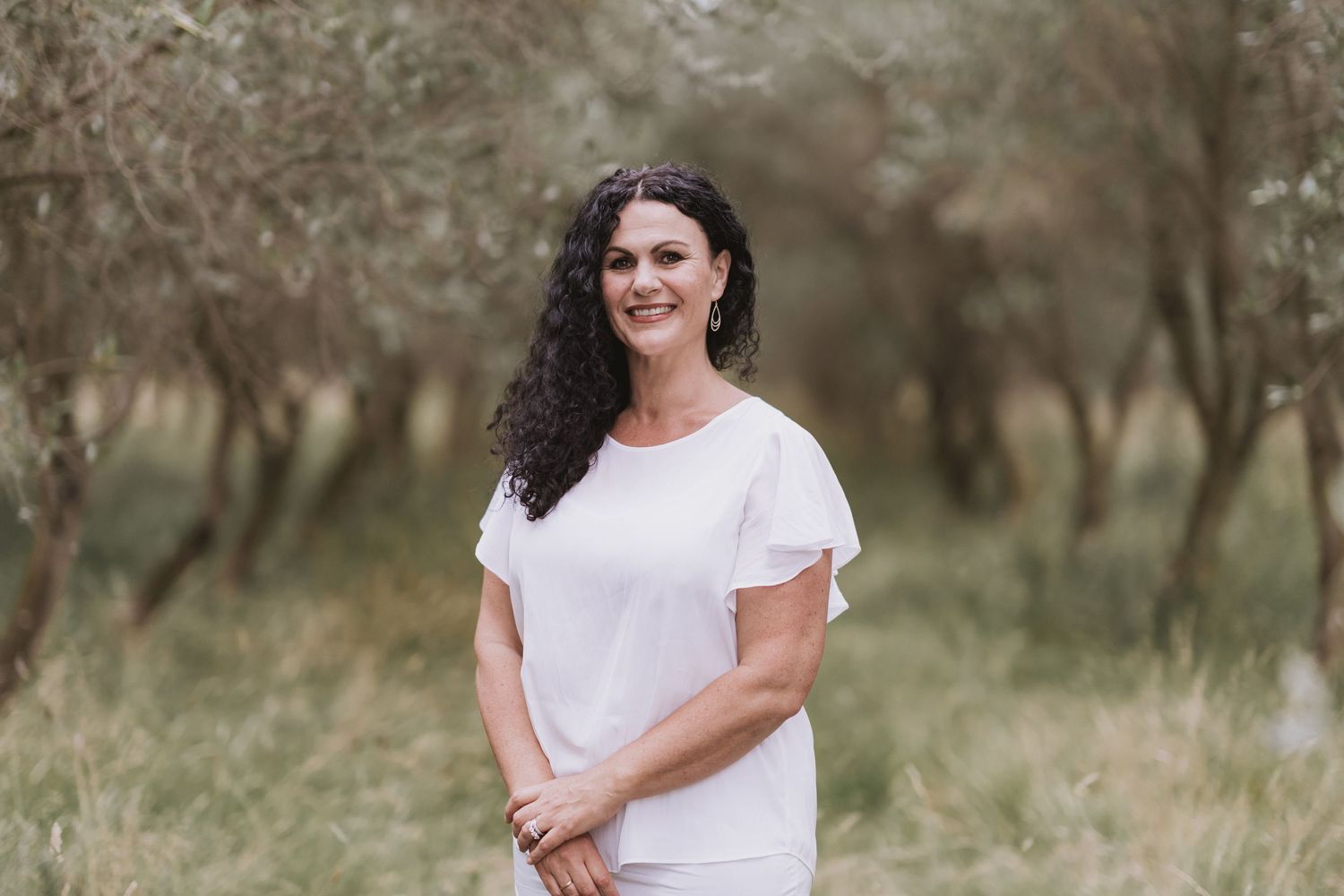A person in a white top smiling while standing in a field with olive trees in the background.